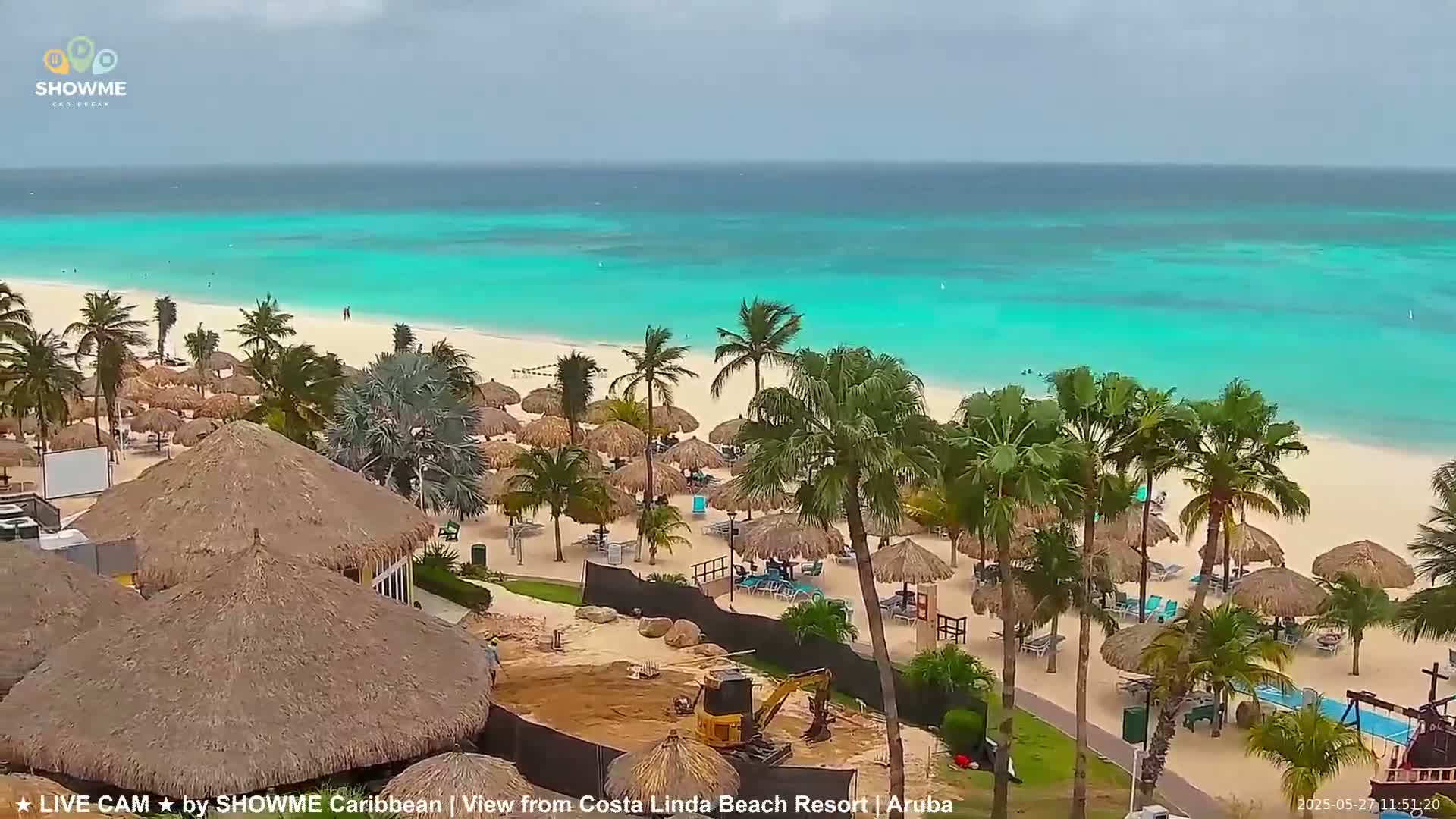 A beach with turquoise water, white sand, palm trees, and thatched-roof umbrellas under an overcast sky.