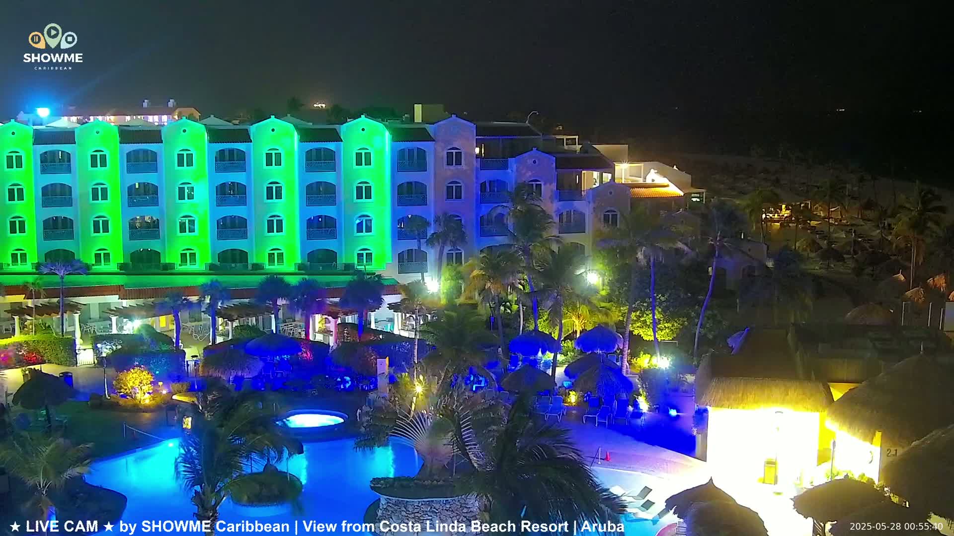A brightly lit resort at night, featuring a multi-story building with green lights, a swimming pool, and palm trees surrounding the property.