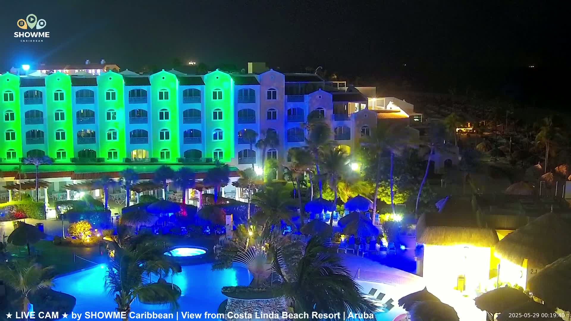 A brightly lit resort at night, featuring a multi-story building with green and blue lights illuminating its exterior, a swimming pool, and surrounding landscaping.