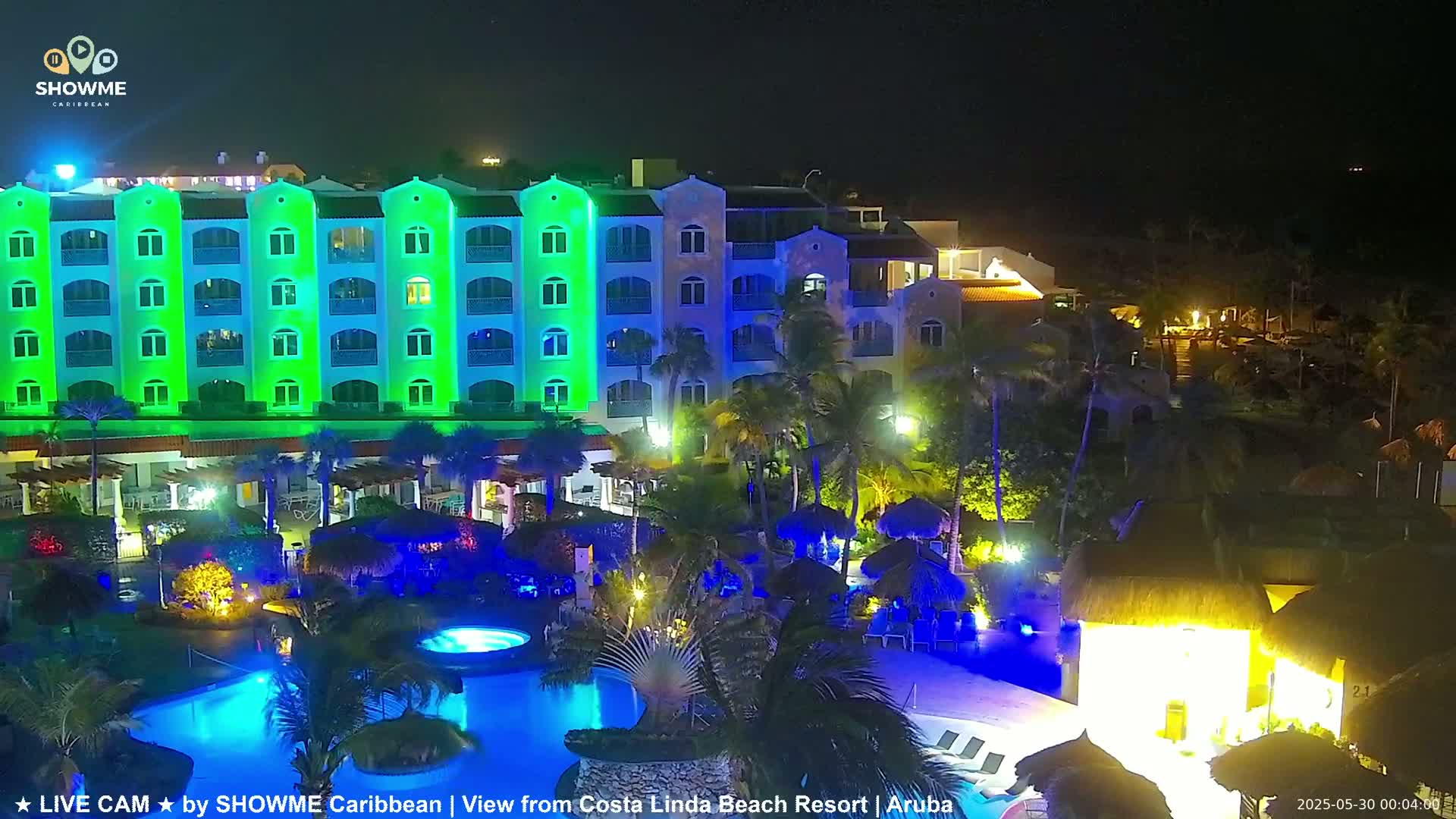 A brightly lit resort at night, featuring a multi-story building with green and blue lighting, a swimming pool, and palm trees.
