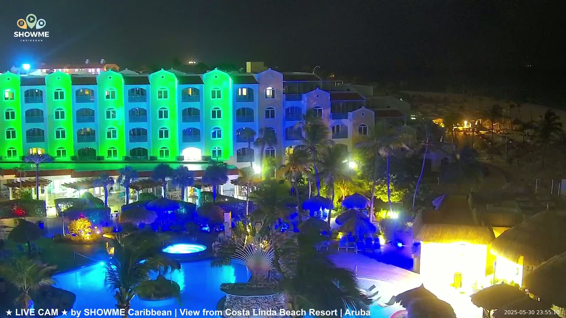 A brightly lit resort at night, featuring a multi-story building with green accent lighting, surrounding pools with blue lighting, and palm trees.