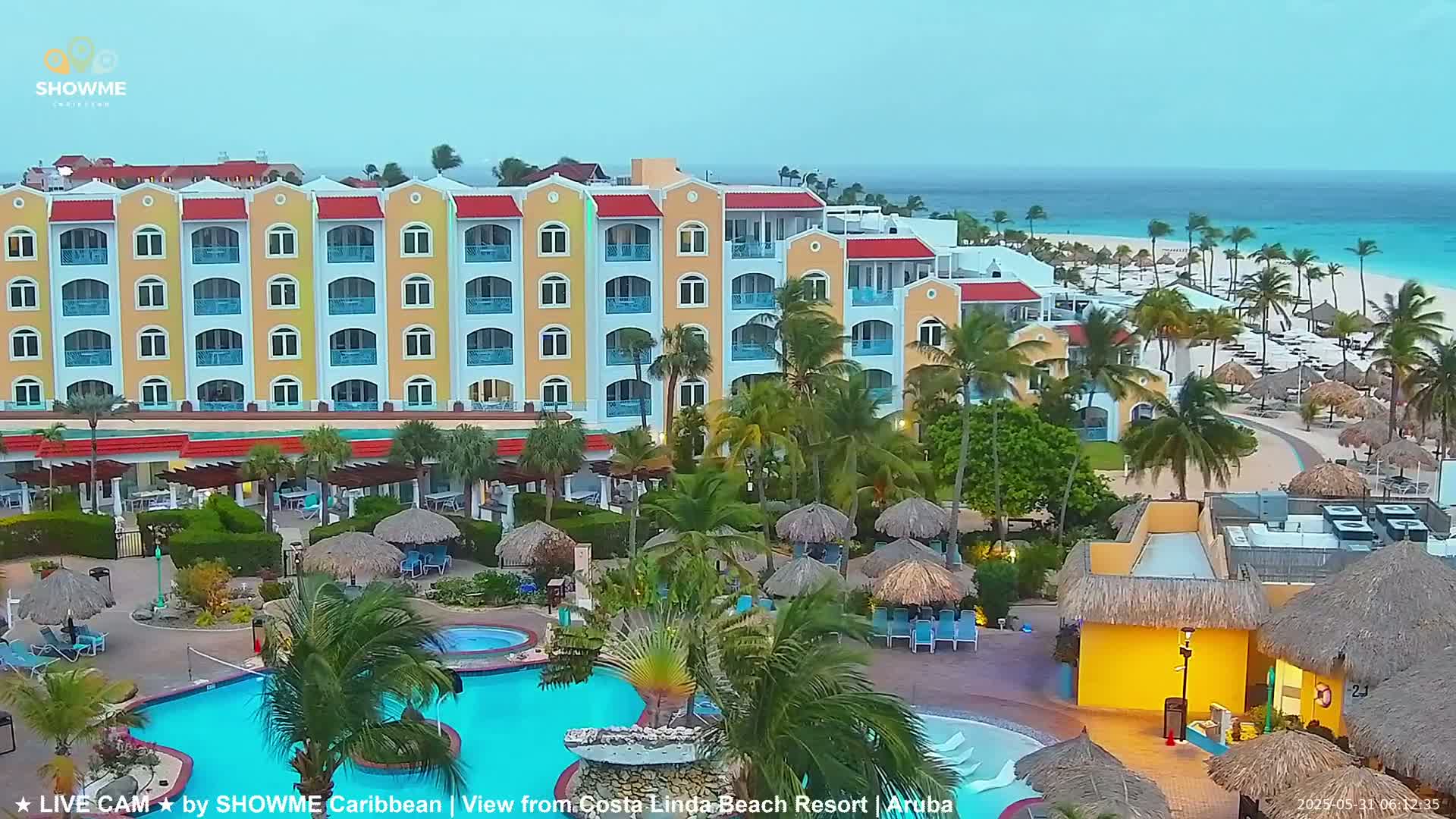 A large resort with a swimming pool and thatch-roofed cabanas is situated near a beach under a partly cloudy sky.