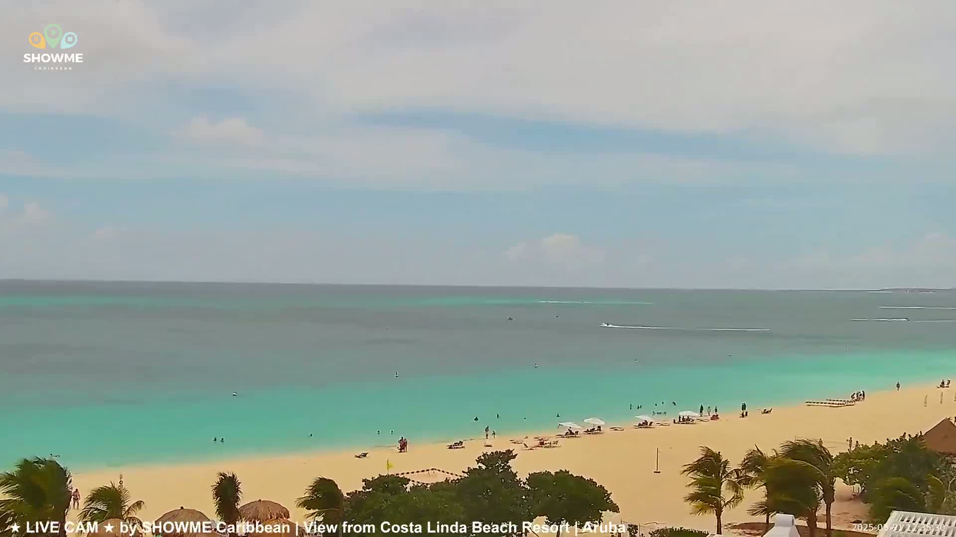 A sandy beach with turquoise water and a few people on it is viewed under a partly cloudy sky.