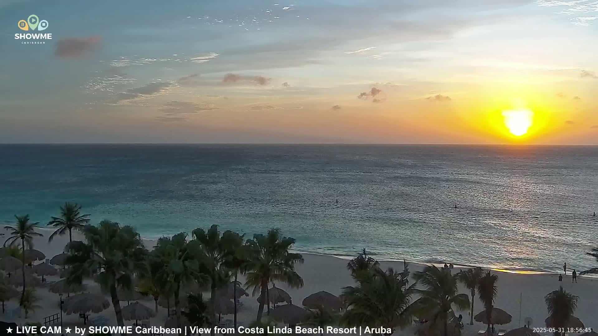 A beach at sunset, with palm trees and thatched umbrellas, overlooks a calm ocean under a partly cloudy sky.