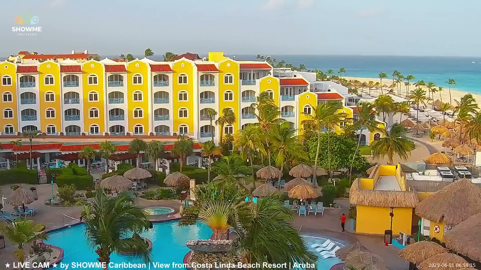A yellow multi-story resort with a pool and palm trees sits on a beach with calm, turquoise water under a clear sky.