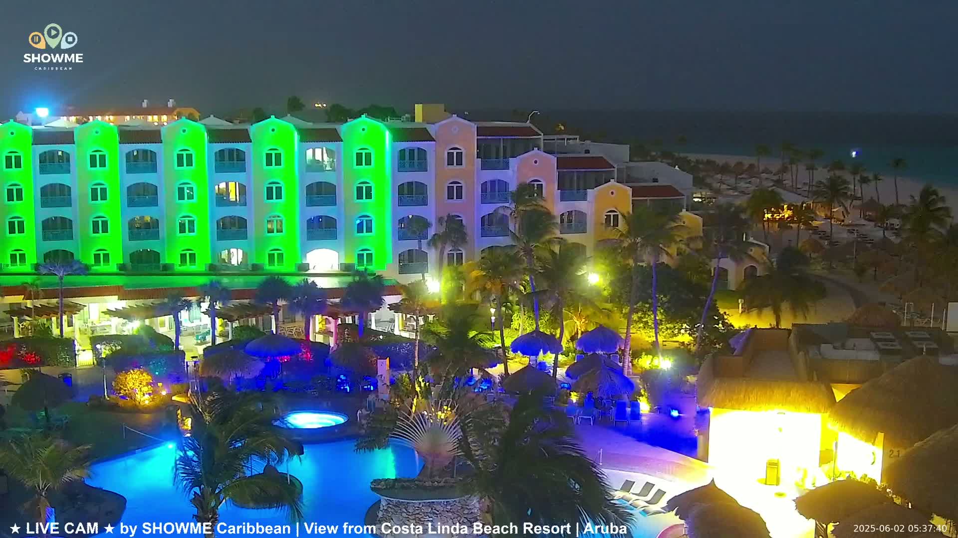A brightly lit resort at night, featuring a large pool and a multi-story building with green lighting, overlooks a beach under a dark sky.