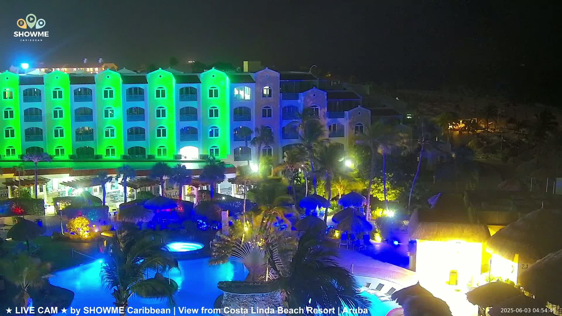 A brightly lit resort at night features a large multi-story building, swimming pools, and tropical landscaping.