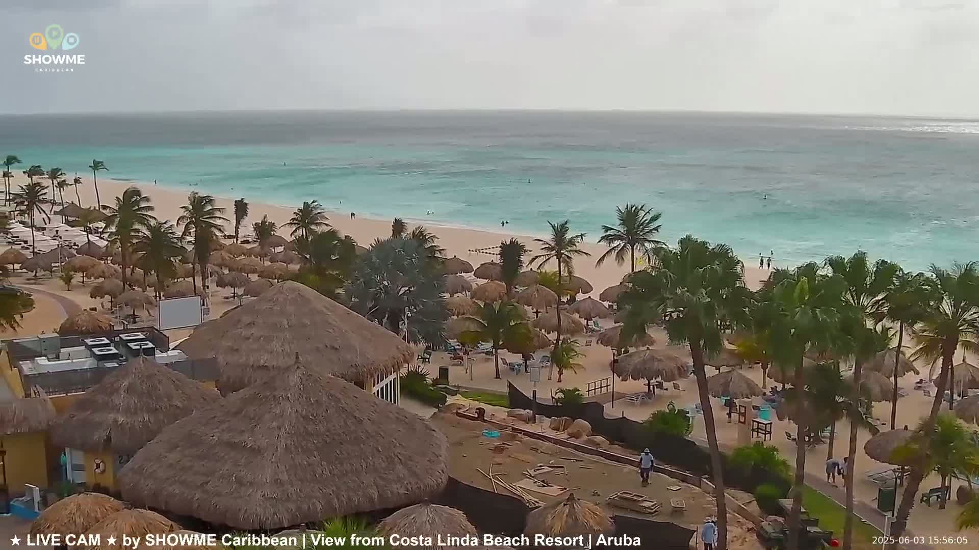 An overcast day on a tropical beach with turquoise water, palm trees, and numerous thatched-roof structures.