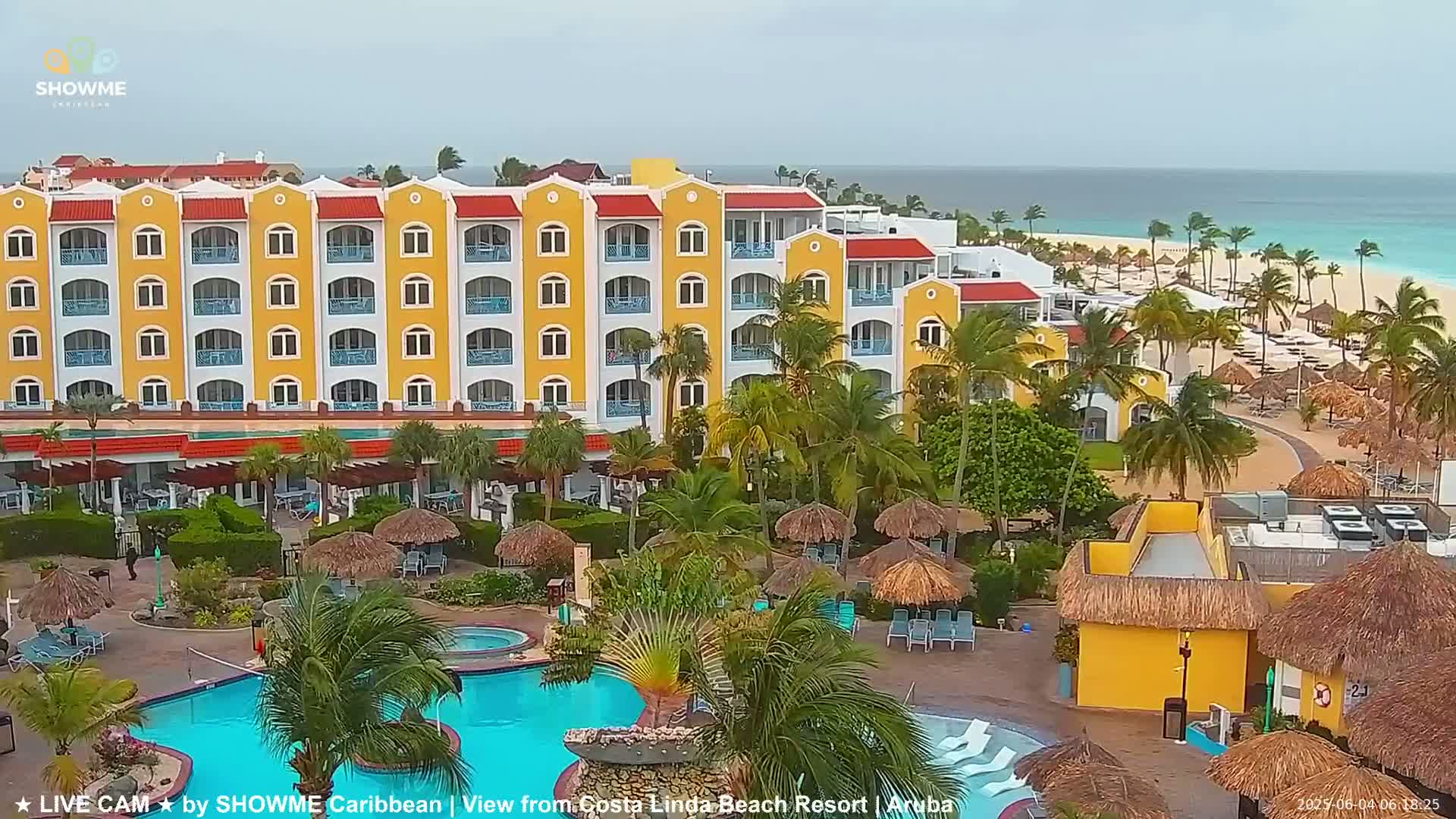 A yellow resort with a swimming pool and palm trees sits on a beach with turquoise water under overcast skies.