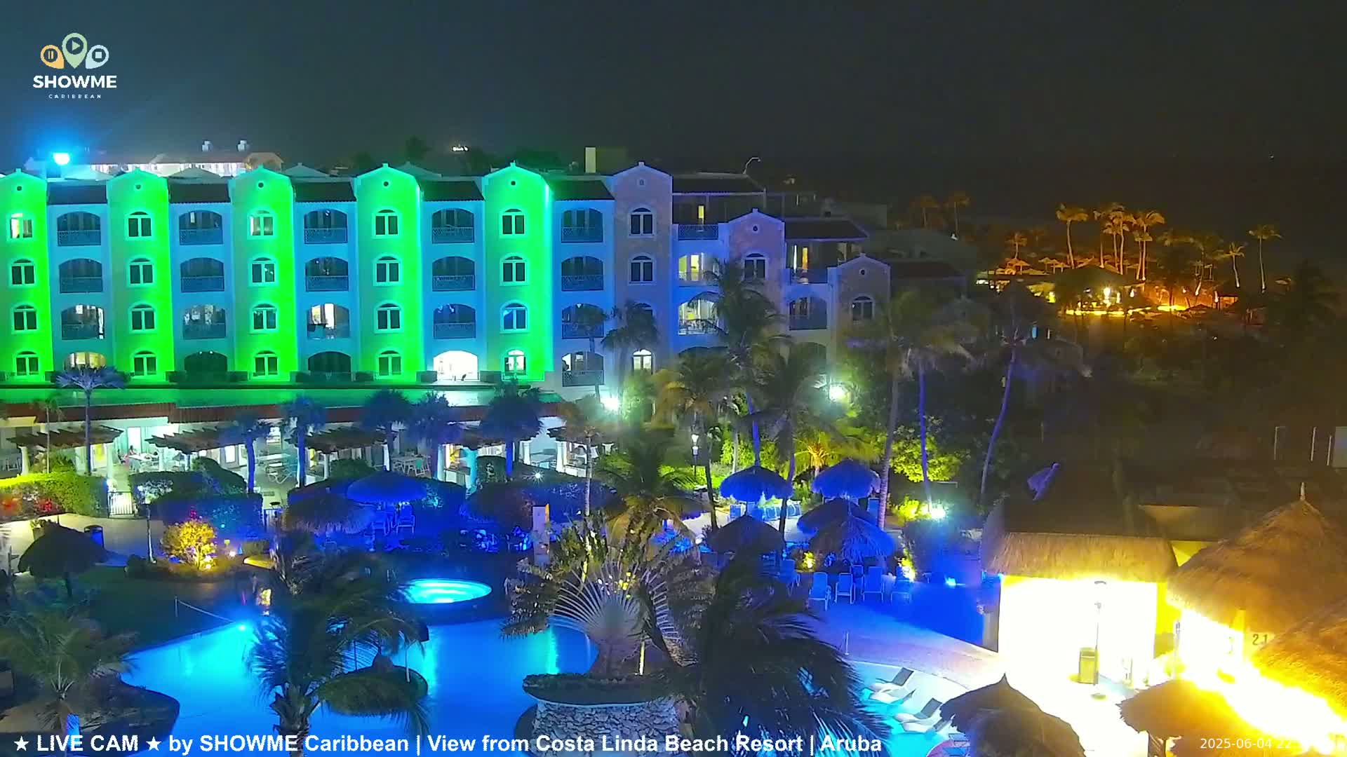 A brightly lit resort at night, featuring a multi-story building with green exterior lighting, a swimming pool, and palm trees.