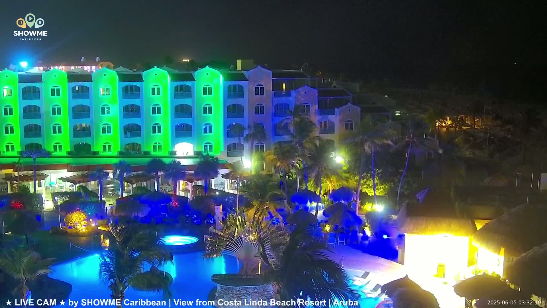 A brightly lit resort at night, featuring a multi-story building with green and blue accent lighting, a swimming pool, and palm trees.