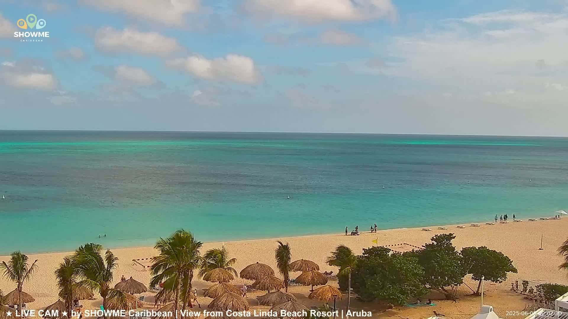 A sandy beach with palm trees and thatched umbrellas stretches along a calm, turquoise ocean under a partly cloudy sky.