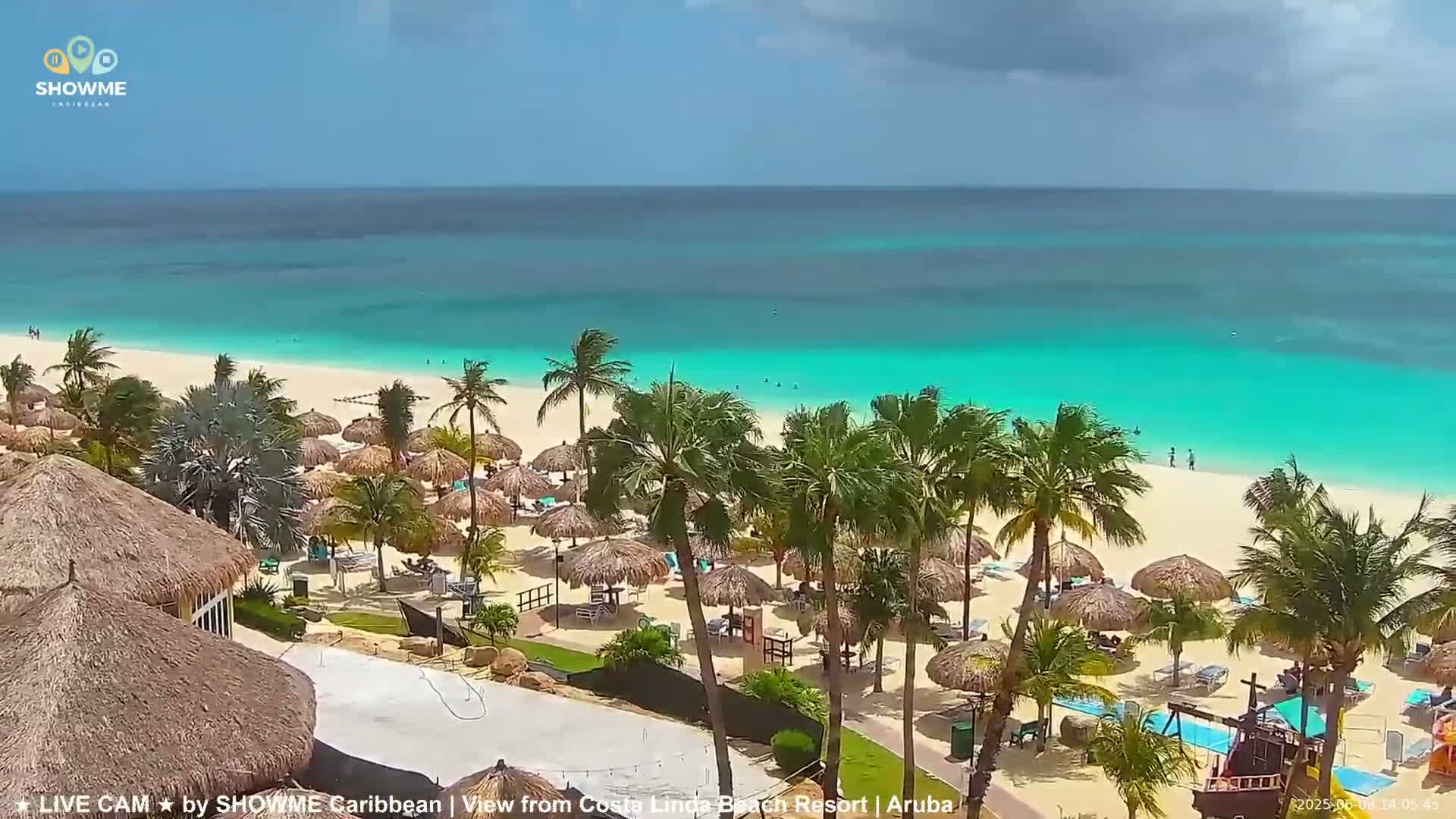 A beach resort with numerous palm trees and thatched umbrellas under partly cloudy skies, overlooking a calm turquoise ocean.