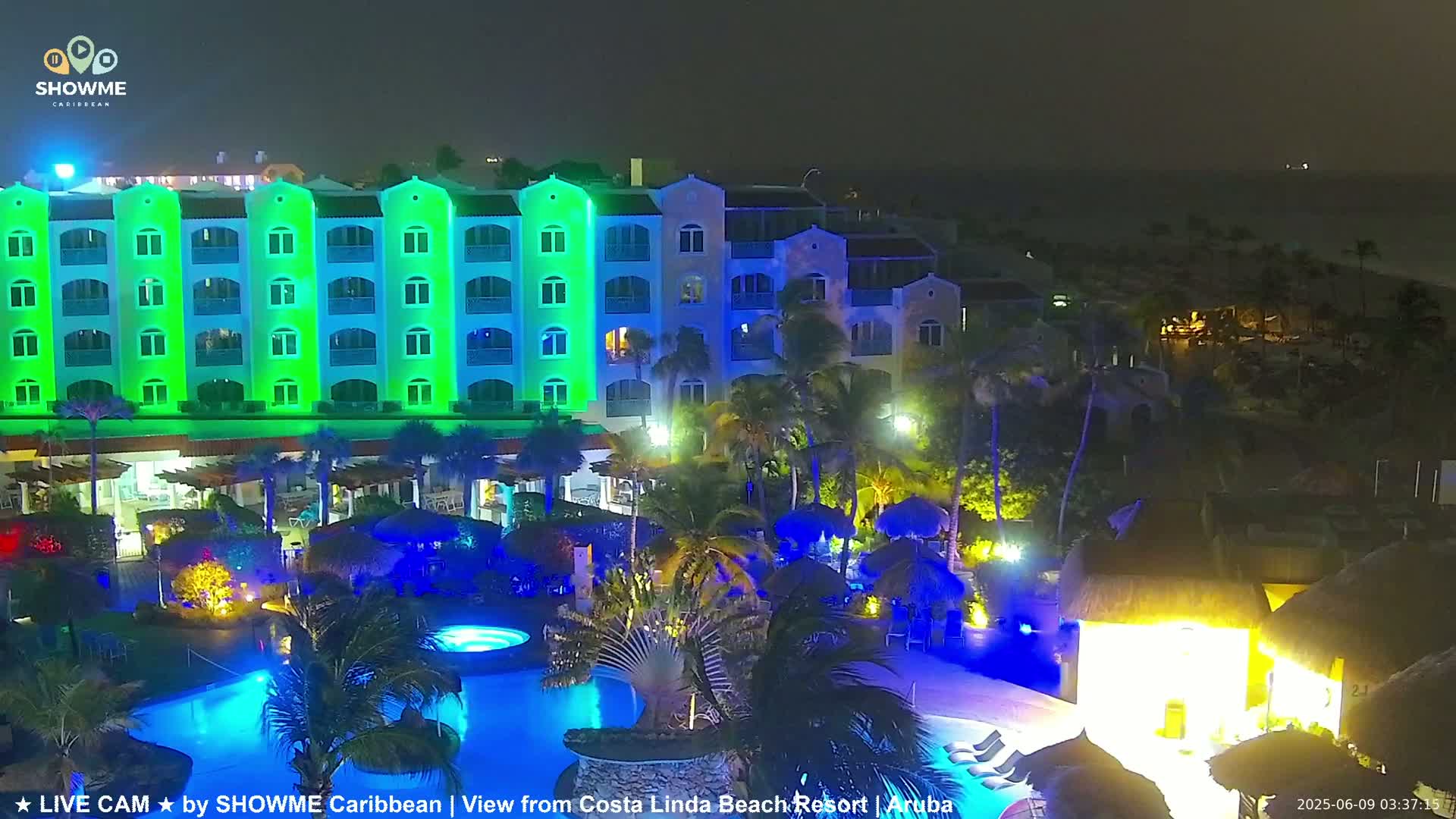 A brightly lit resort at night, featuring a multi-story building with green lights, a swimming pool, and palm trees, overlooks a dark ocean.