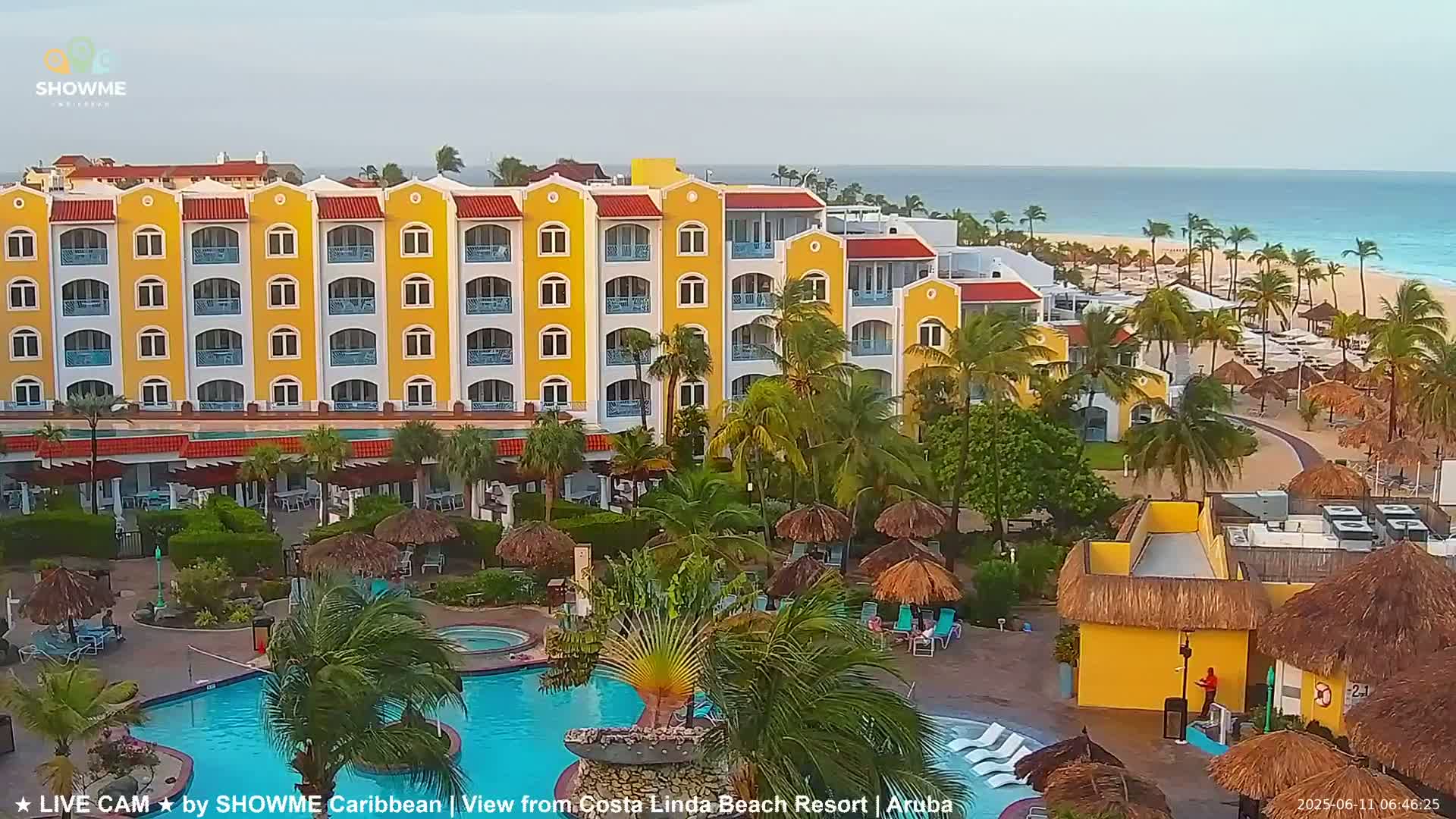 A yellow multi-story resort building with palm trees and a swimming pool is situated near a beach under a partly cloudy sky.