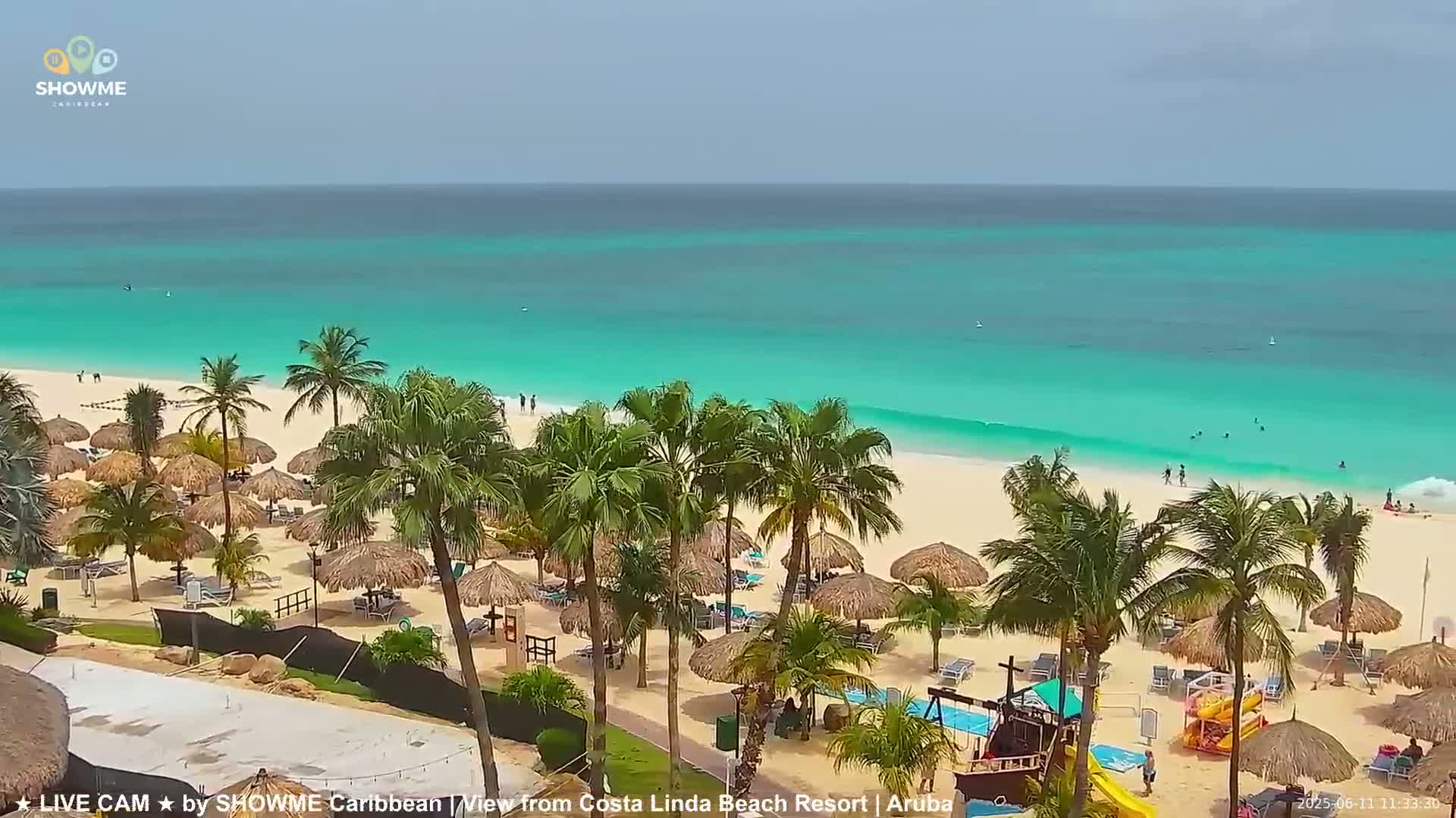 A beach with turquoise water, palm trees, and thatched umbrellas under a partly cloudy sky.
