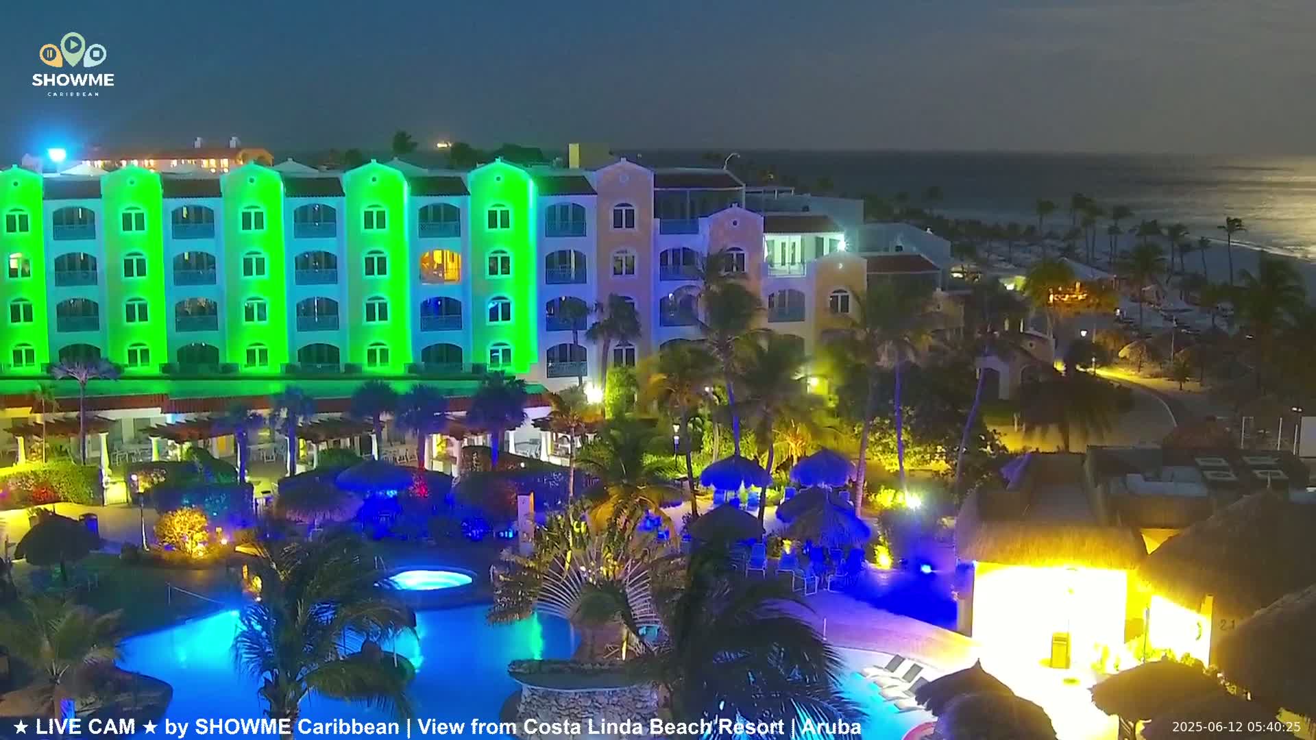 A brightly lit resort at night, featuring a large pool and a multi-story building illuminated in green and blue, overlooks a calm ocean under a dark sky.