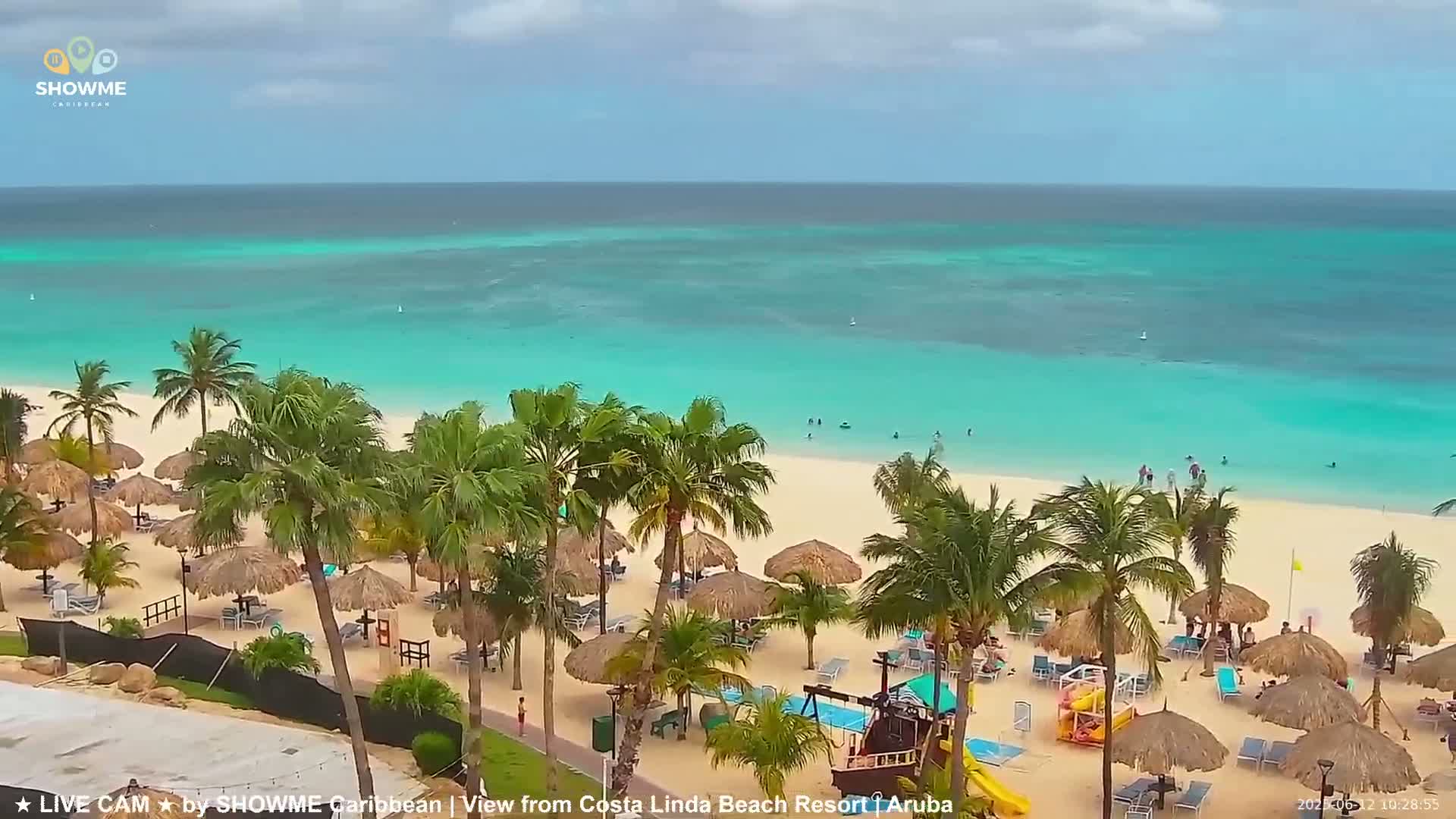 A tropical beach with turquoise water, palm trees, and thatched umbrellas under a partly cloudy sky.