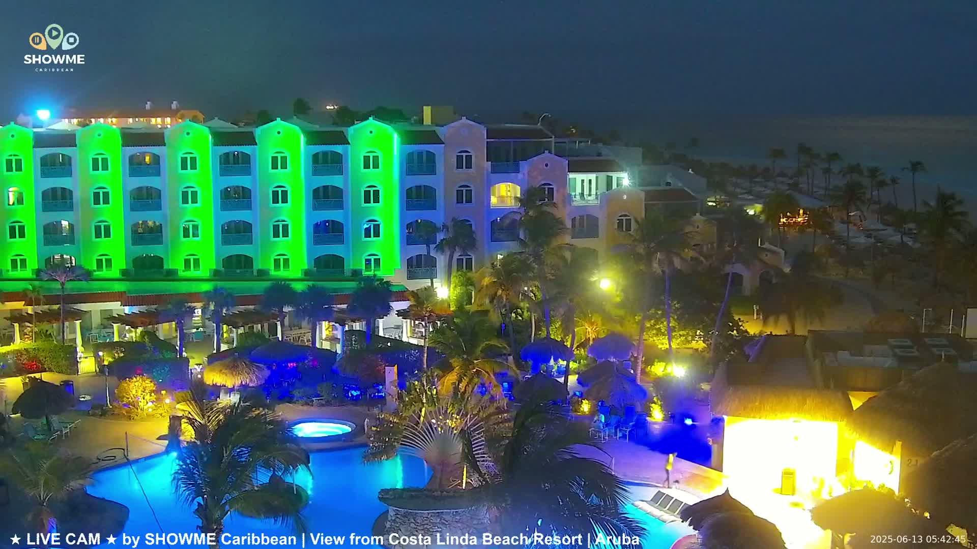 A brightly lit resort at night, featuring a large swimming pool and a multi-story building illuminated in green, under a dark sky.