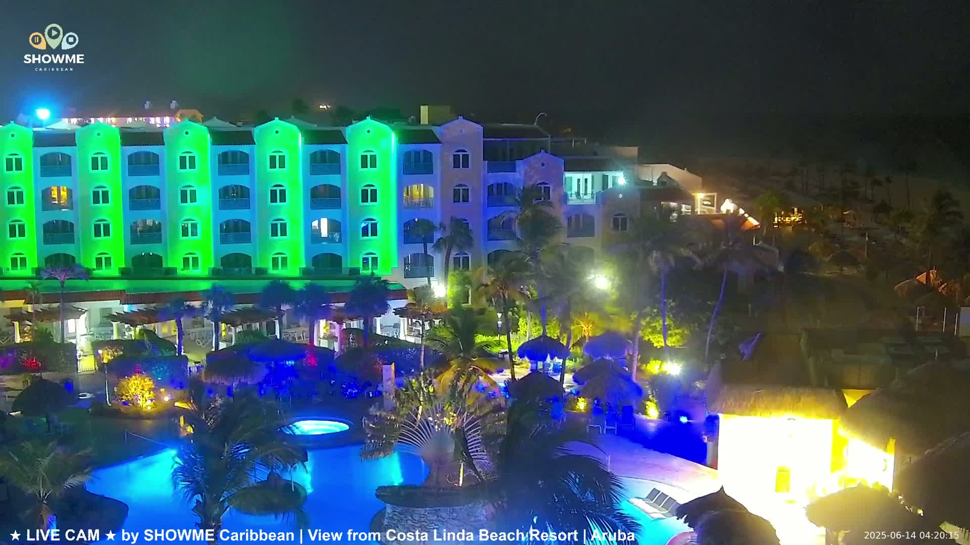 A brightly lit resort at night, featuring a multi-story building illuminated in green and blue, a swimming pool with blue lighting, and surrounding palm trees.