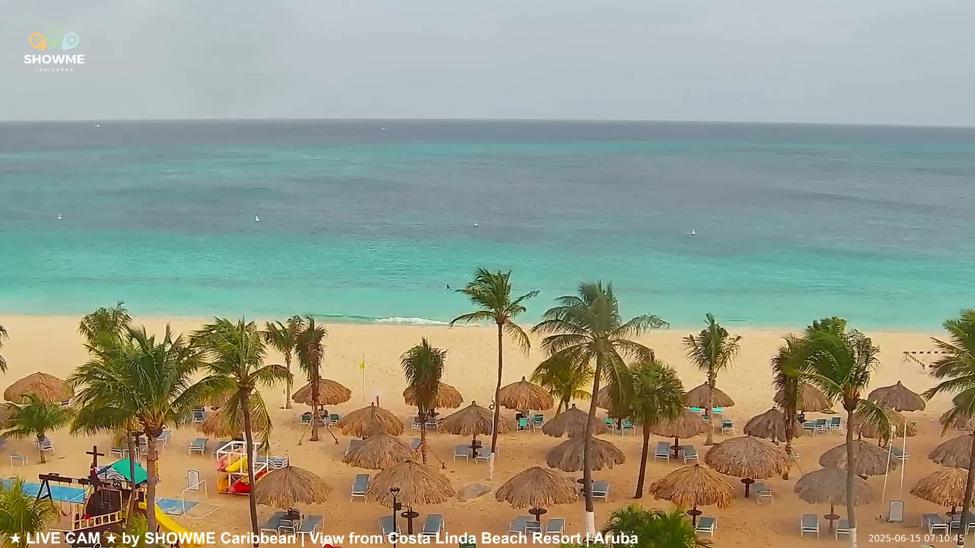 A sandy beach with palm trees and thatched umbrellas is visible under an overcast sky, overlooking a calm turquoise ocean.