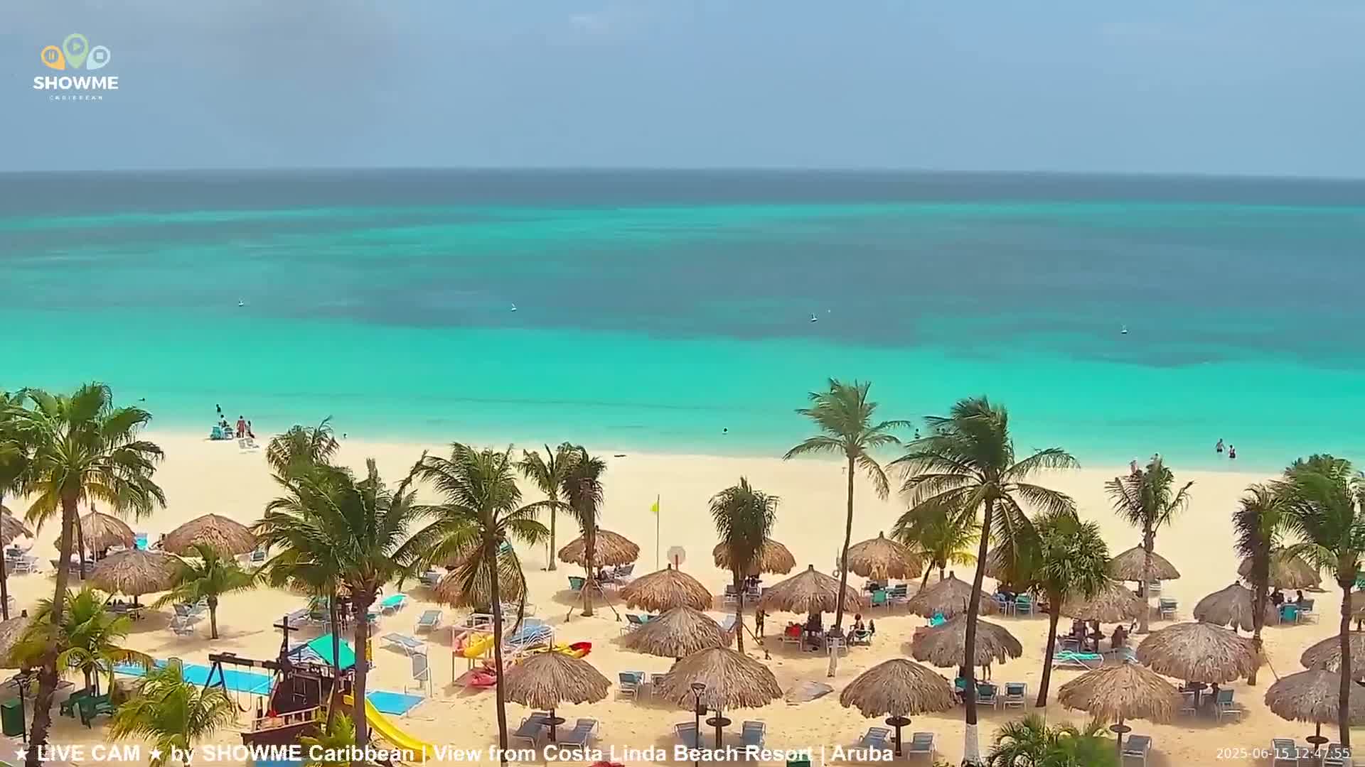 A sandy beach with numerous thatched umbrellas, palm trees, and beachgoers is shown, overlooking a calm, turquoise ocean under a bright, sunny sky.