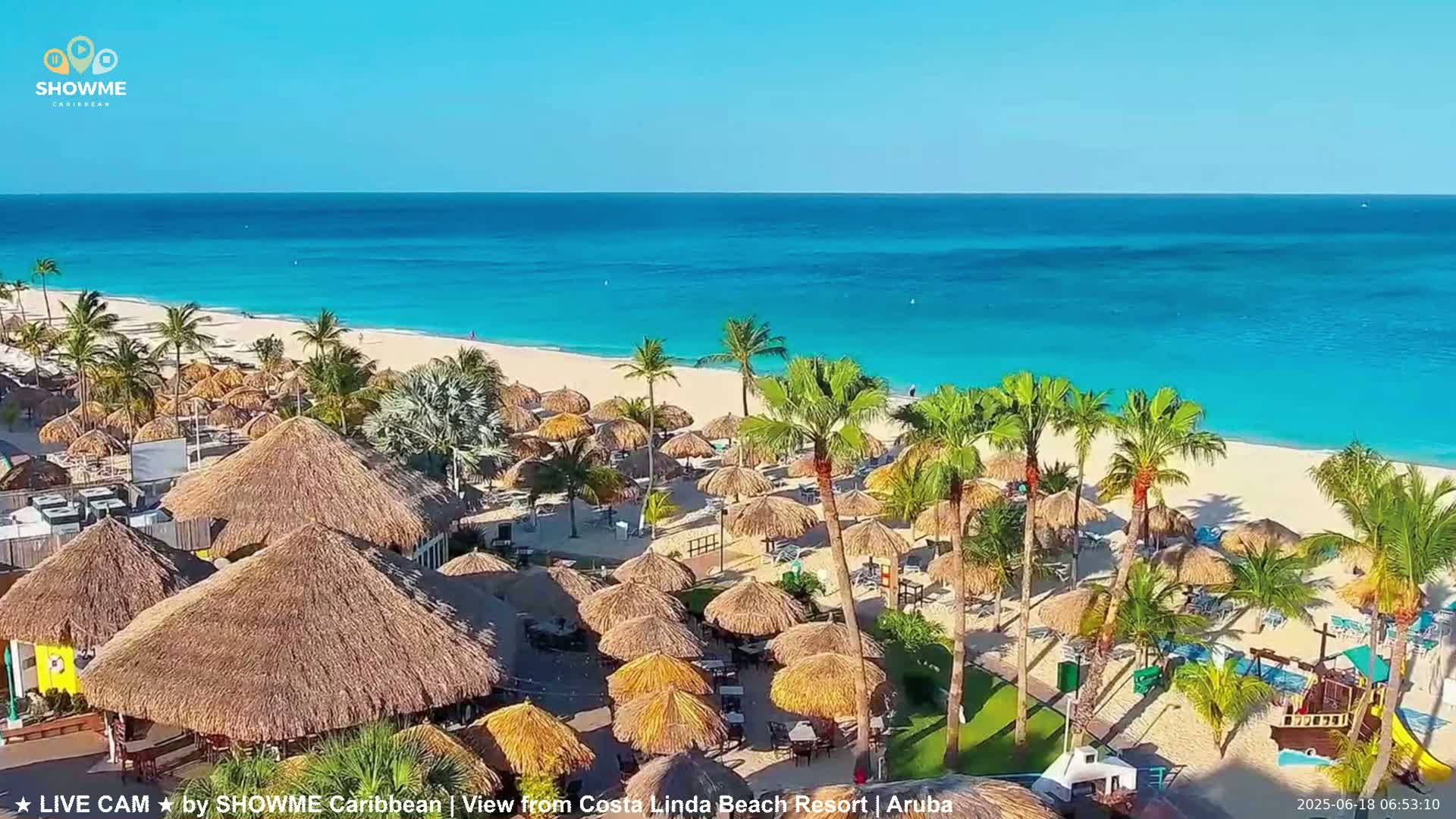 An aerial view shows a beach resort with numerous thatched-roof umbrellas lining a pristine white sand beach next to a turquoise ocean under a clear blue sky.