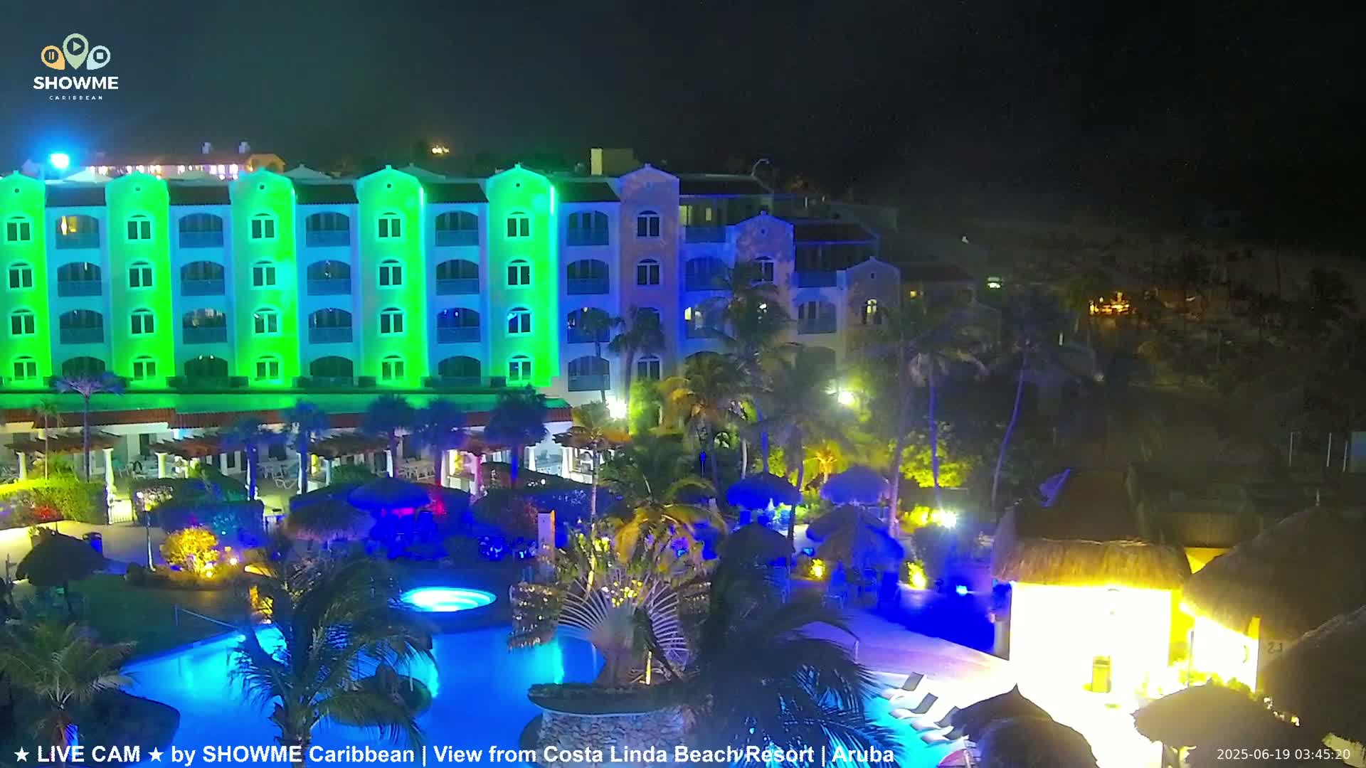 A brightly lit resort at night with a swimming pool and palm trees surrounding a multi-story building.