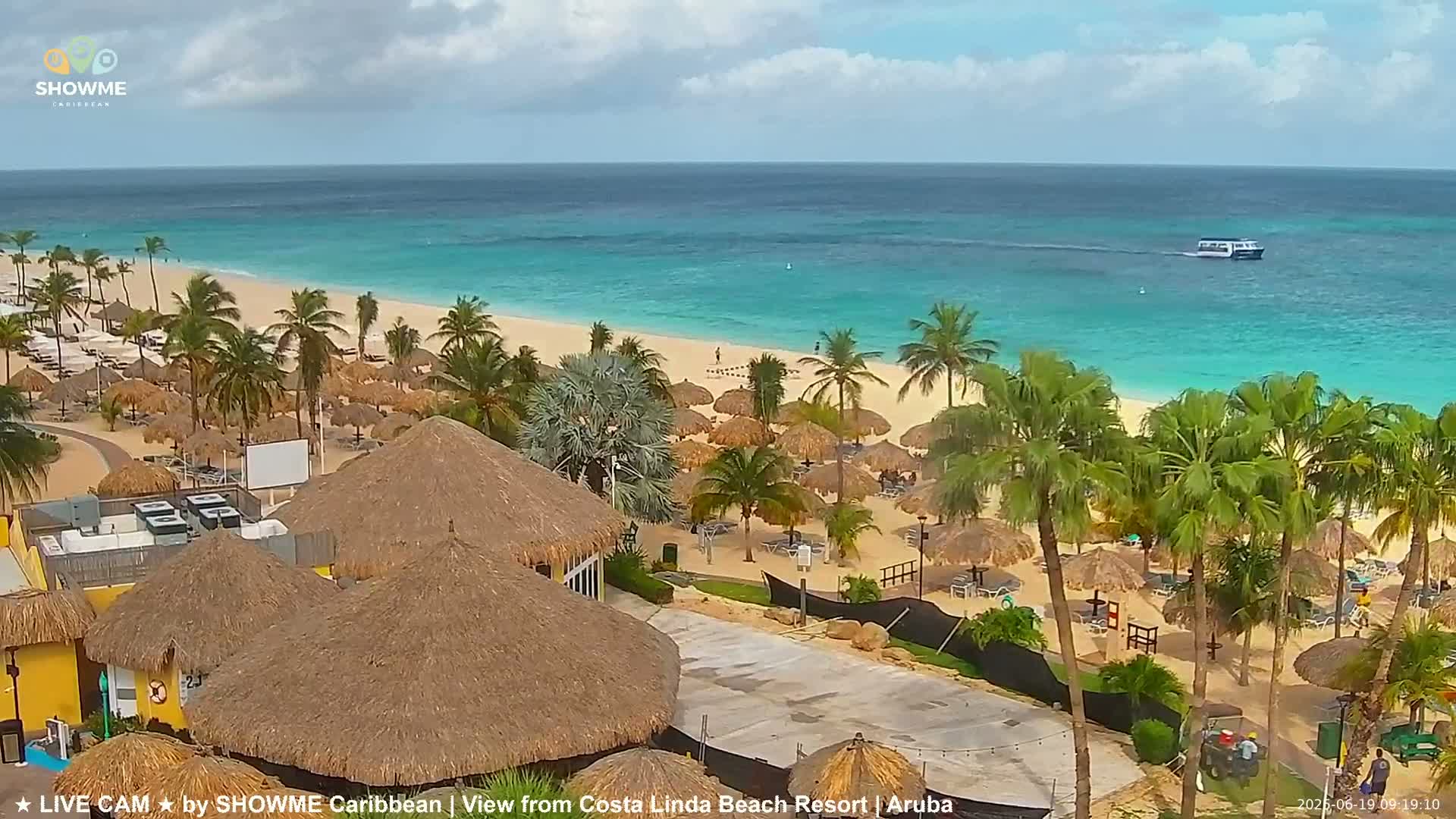 A beach resort with numerous thatched-roof structures overlooks a calm, turquoise ocean under partly cloudy skies.