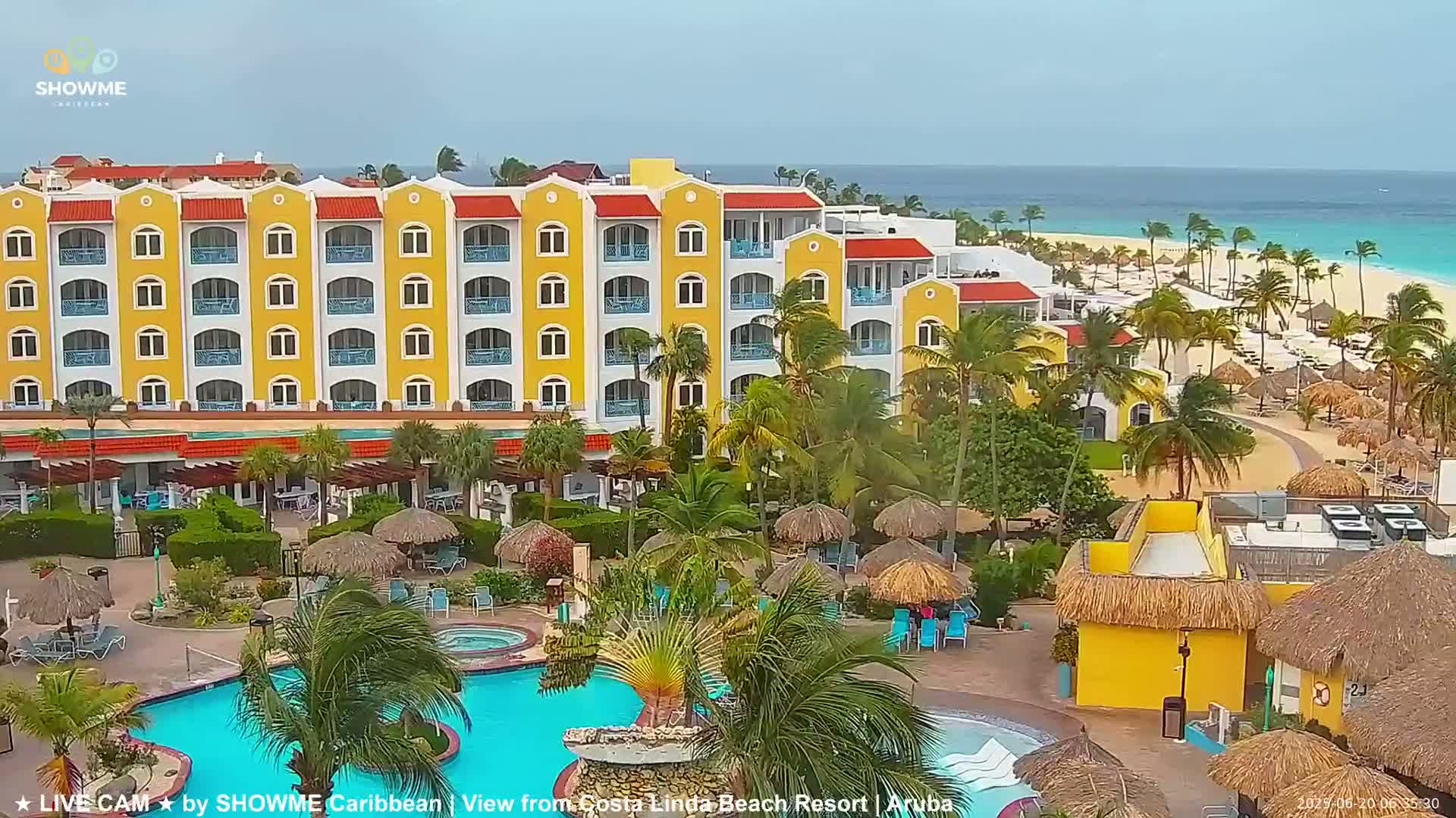 A yellow resort building with palm trees and a pool is situated near a beach under partly cloudy skies.