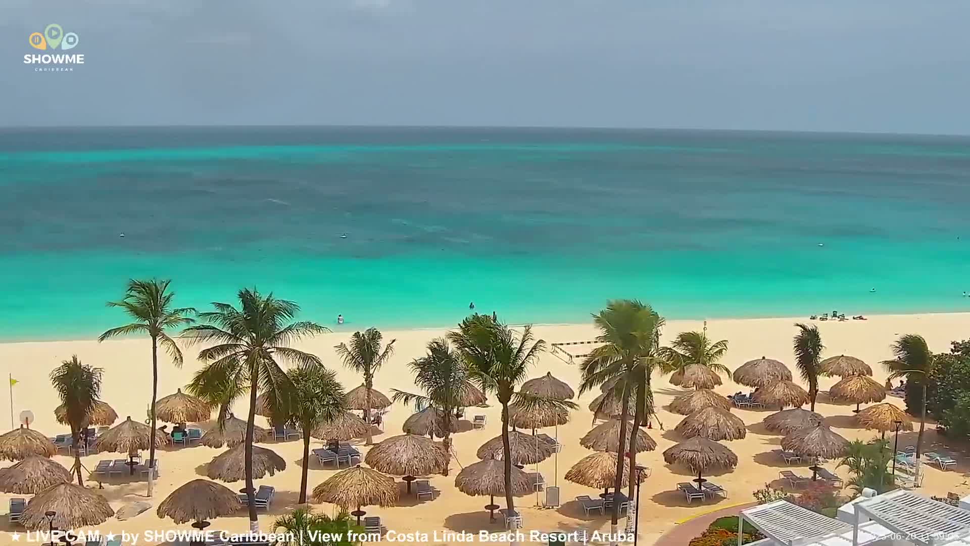A pristine white-sand beach lined with thatched umbrellas and palm trees meets a vibrant turquoise ocean under a mostly clear sky.