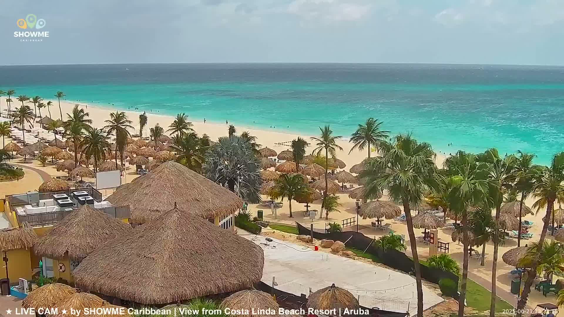 A beach resort with thatched-roof structures overlooks a calm, turquoise ocean under a partly cloudy sky.