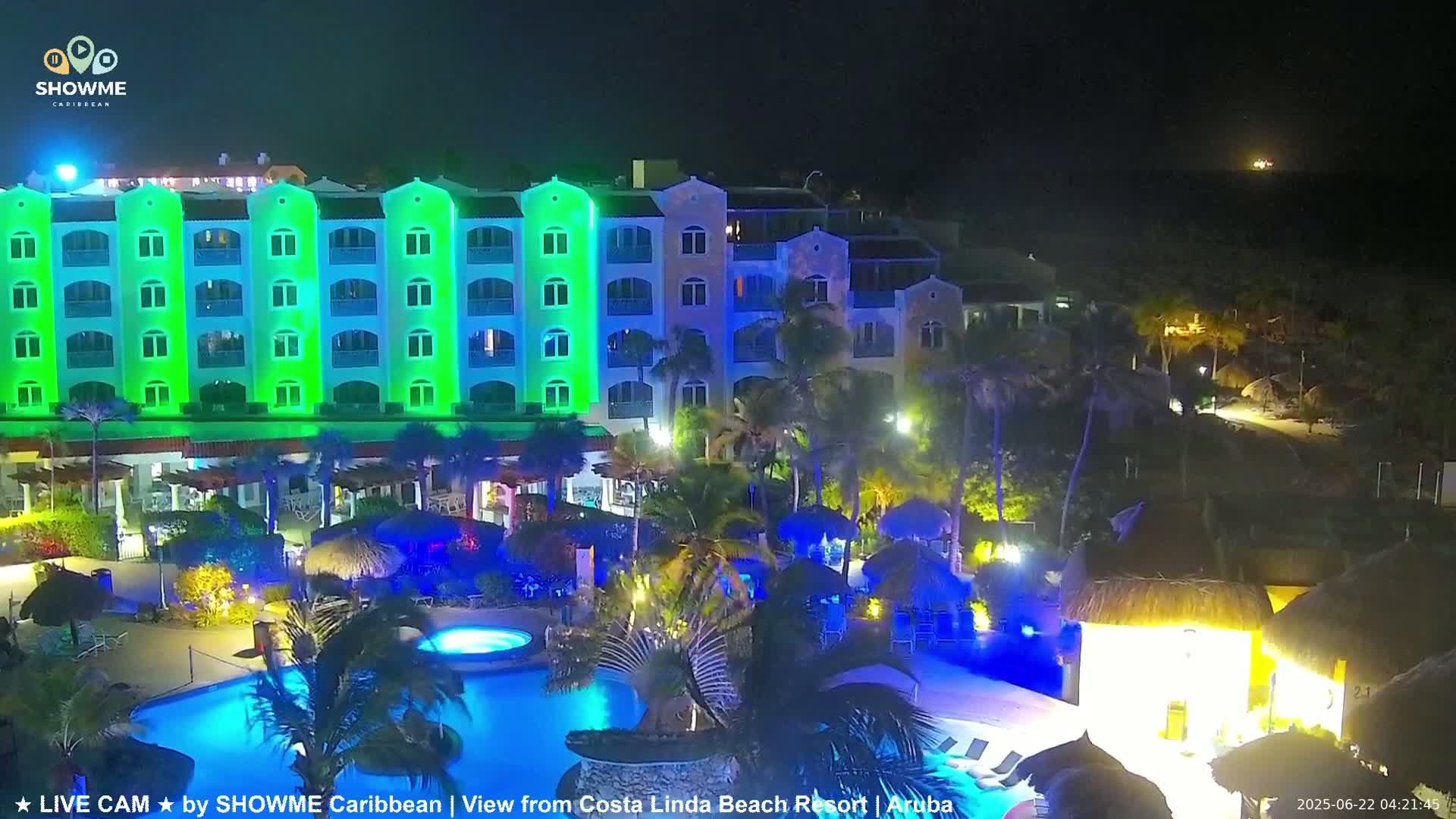 A brightly lit resort at night, featuring a large swimming pool and a multi-story building illuminated in green.