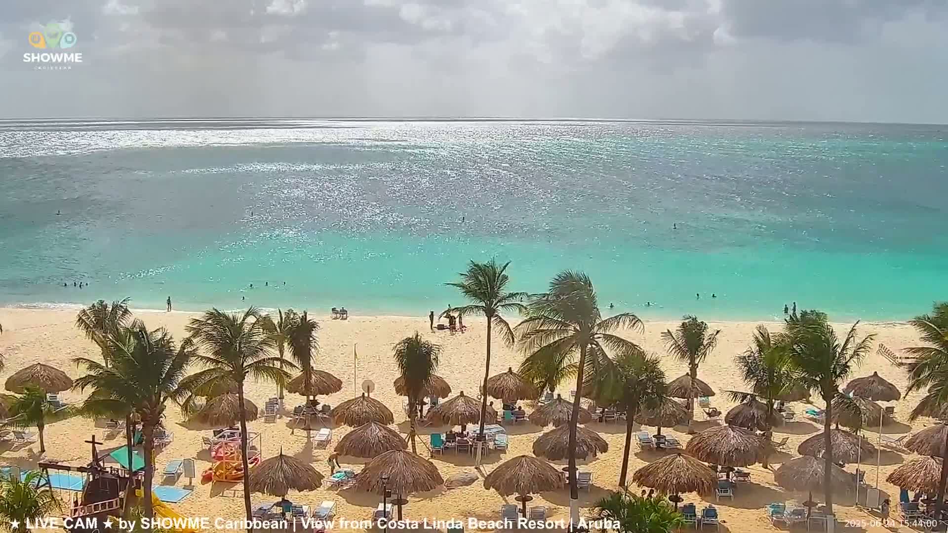 A sandy beach with turquoise water, palm trees, and thatched umbrellas is visible under a partly cloudy sky.