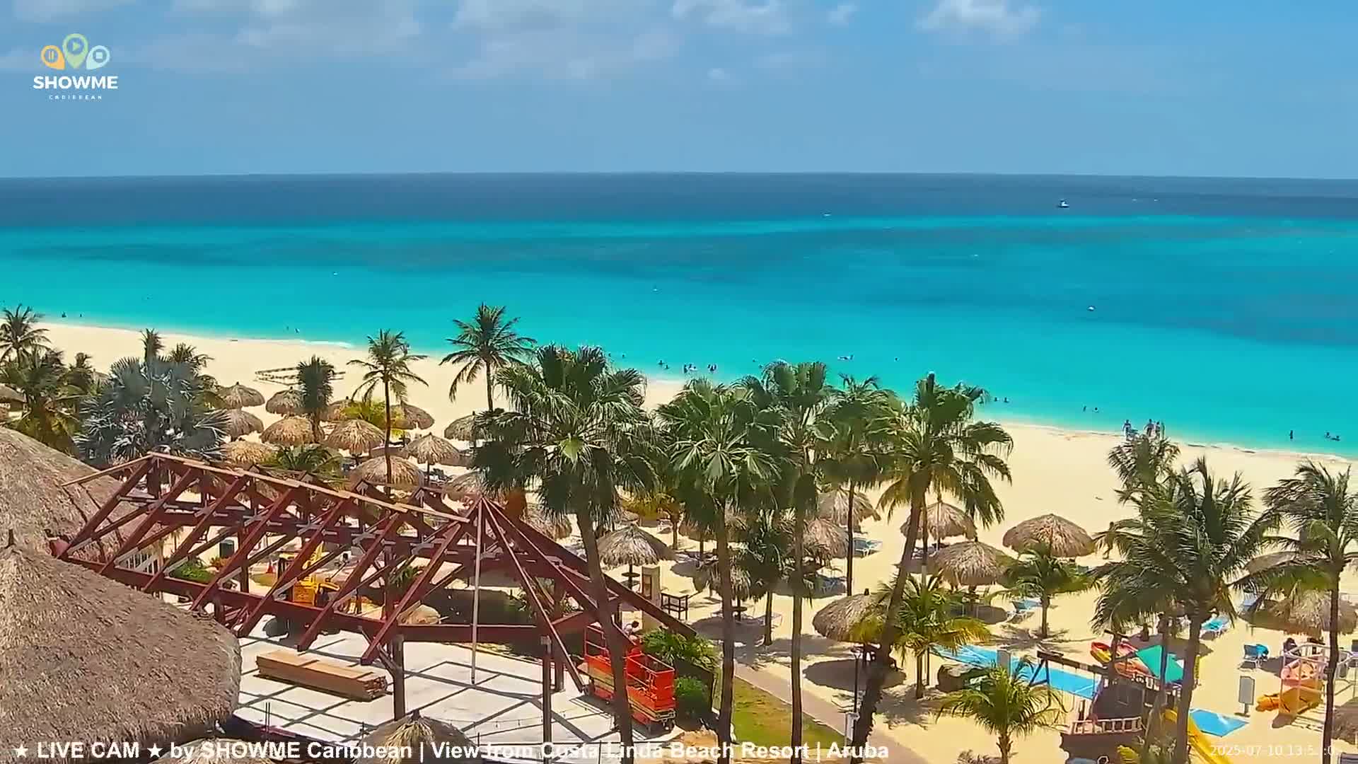 A sunny day at a beach resort shows a turquoise ocean, white sand, palm trees, beach umbrellas, and a partially constructed structure.