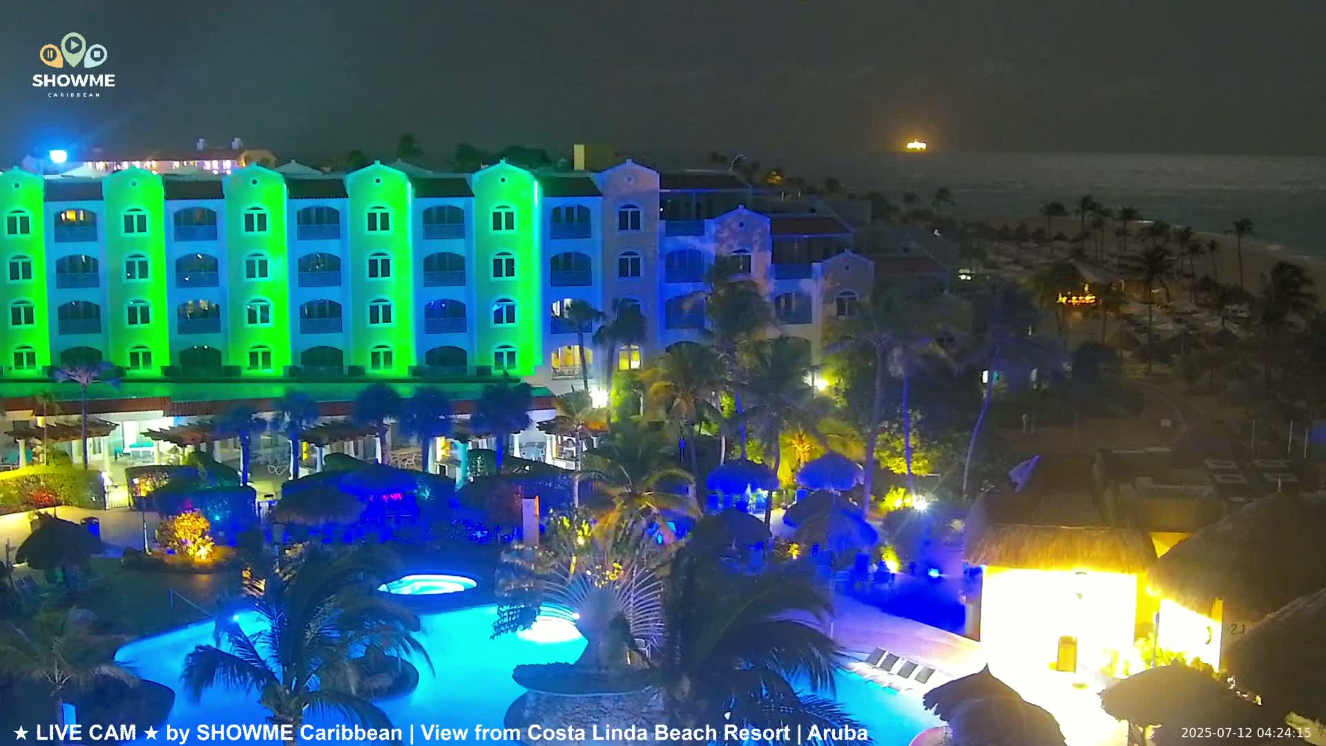 A brightly lit resort at night, featuring a multi-story building with pools and palm trees, overlooks a calm ocean under a dark sky.