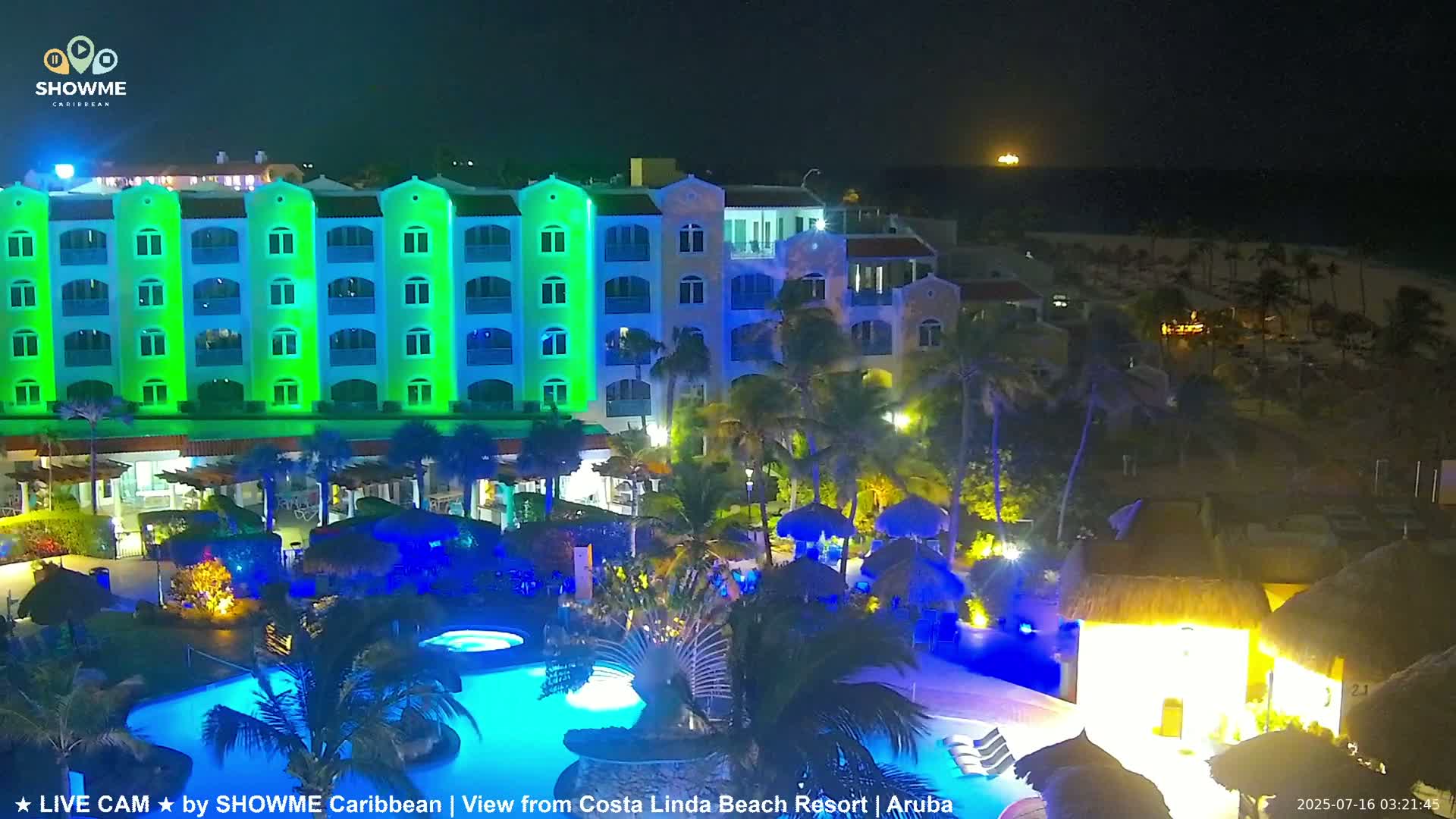 A brightly lit resort at night, featuring a multi-story building with green and blue lighting, surrounding pools illuminated in blue, and palm trees under a clear, dark sky.