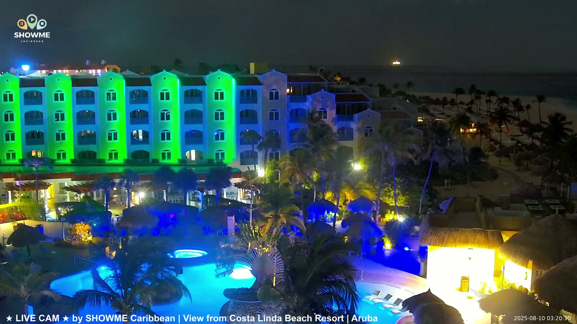 A brightly lit resort at night, featuring a multi-story building with green and blue lights, pools illuminated in blue, and palm trees under a dark sky.