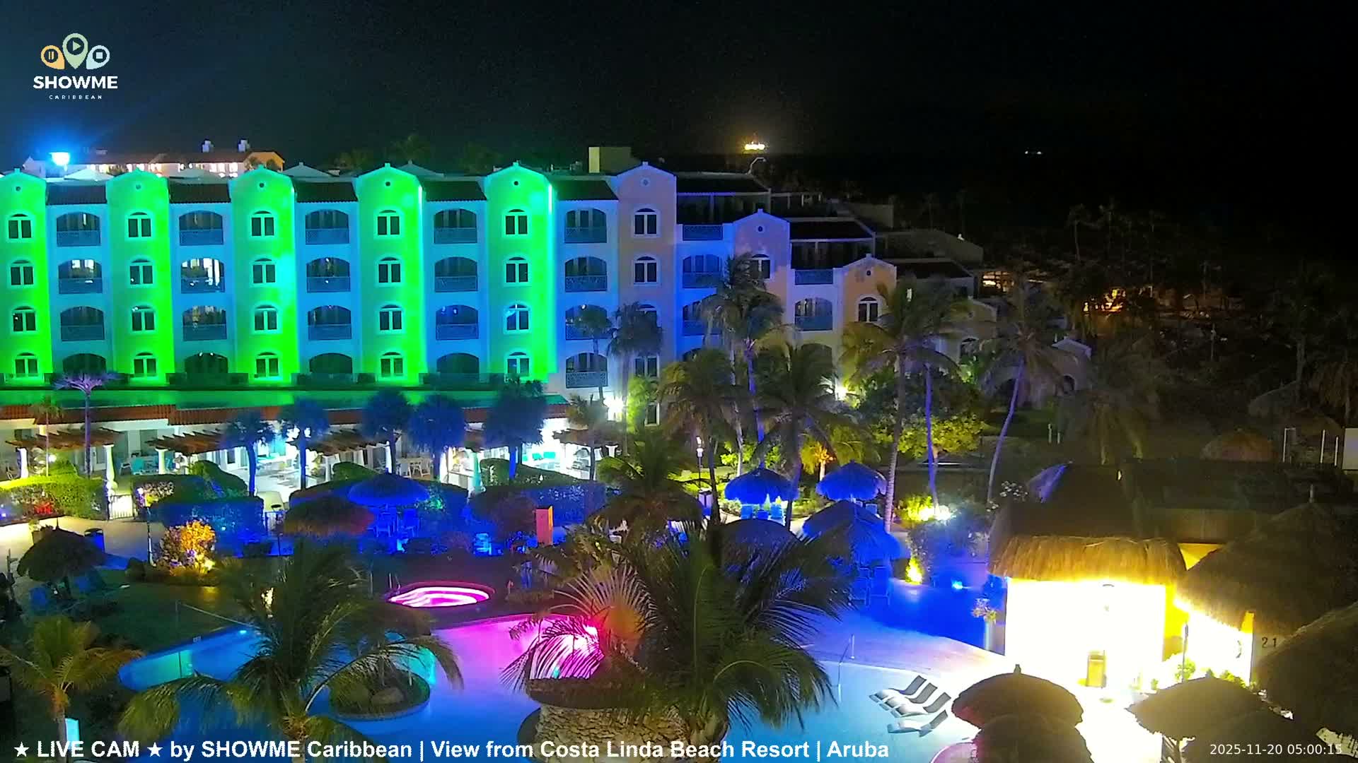 The image captures a vibrant night scene at a tropical resort, featuring a multi-story building illuminated in green and blue, lush palm trees, swimming pools, a hot tub with pink lighting, and thatched-roof structures, all under a clear dark sky.