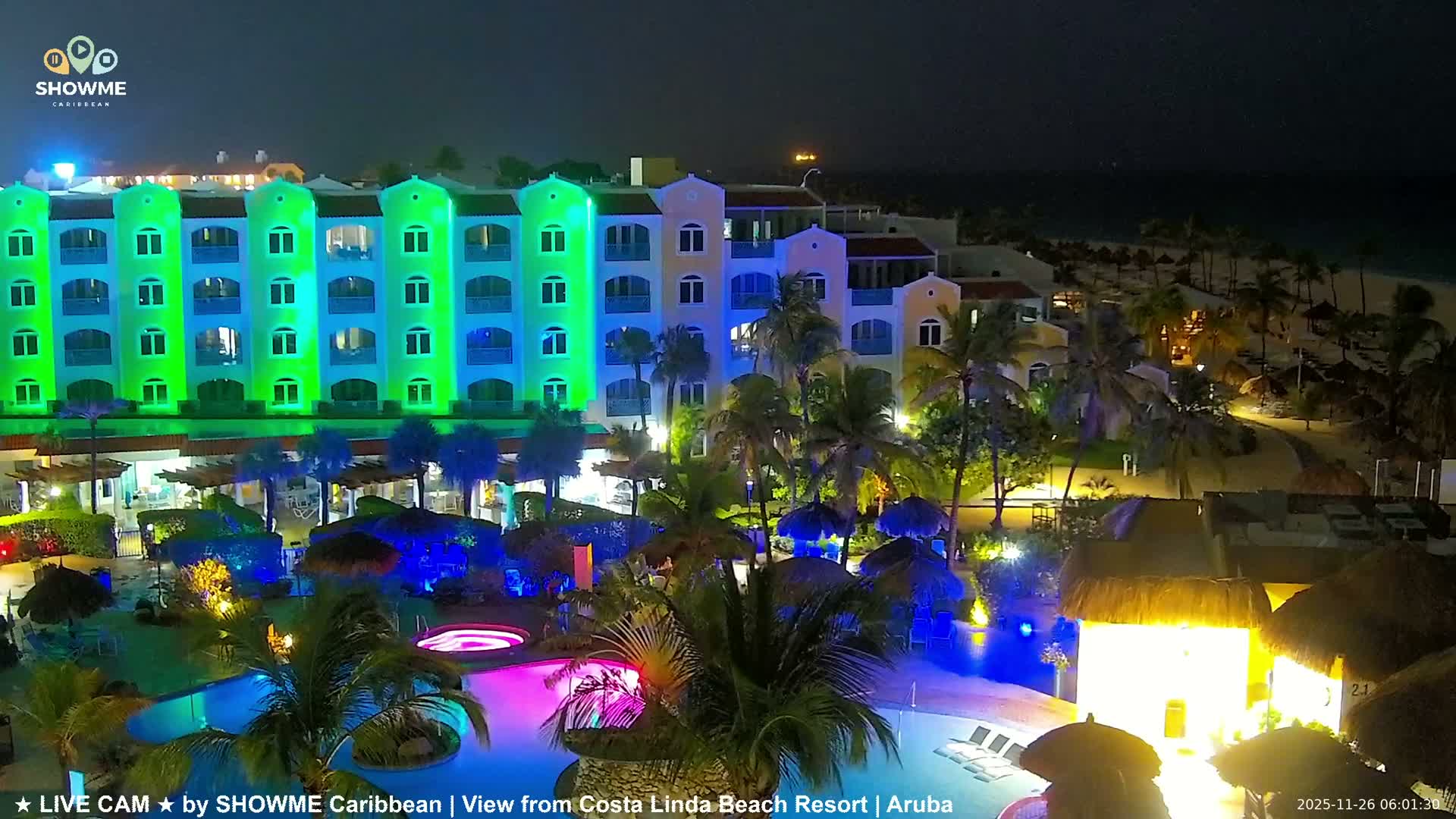 A vibrant nighttime view reveals a tropical resort with multi-colored buildings illuminated in green and blue, lush palm trees, glowing pools, and a sandy beach stretching towards the dark ocean under a clear sky.