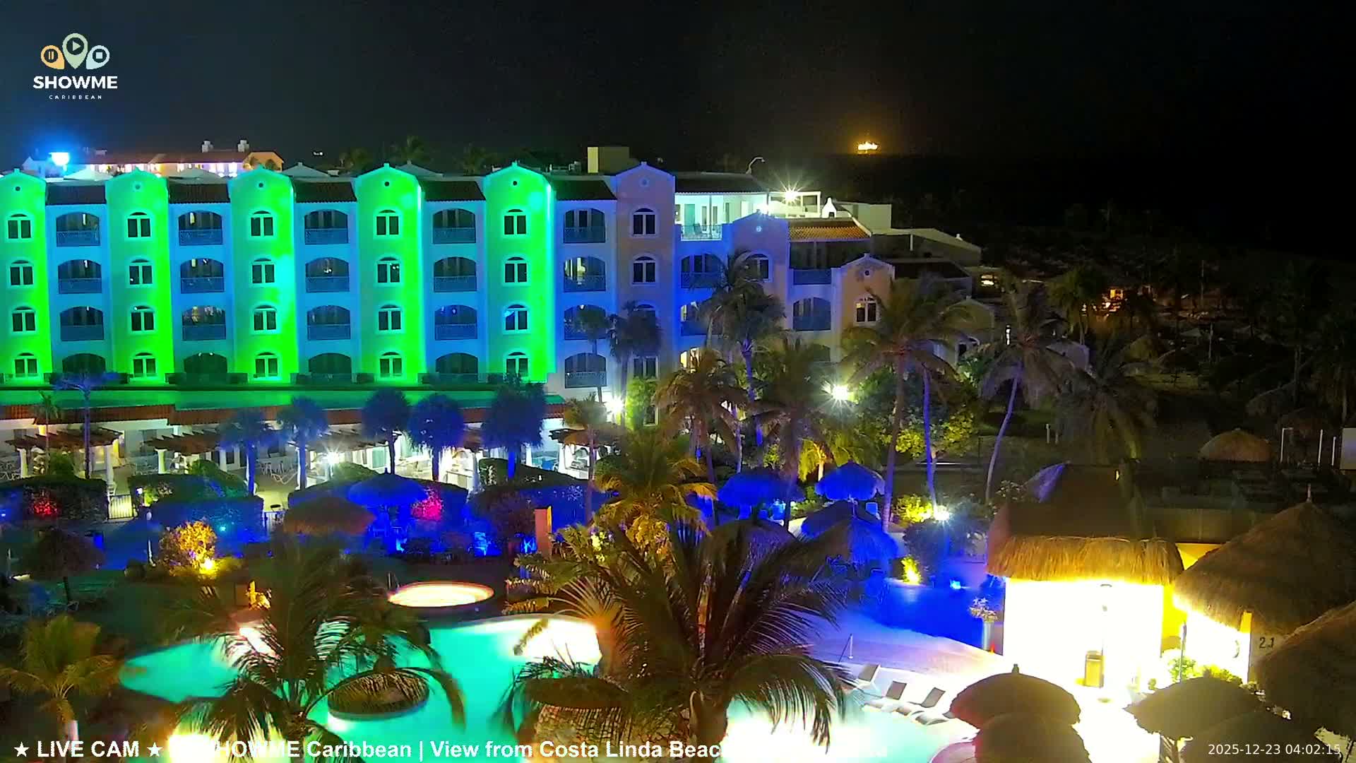 A vibrant resort complex is seen at night, featuring multi-story buildings illuminated in alternating green and blue lights, tropical palm trees, and sparkling blue swimming pools, all under a clear dark sky.