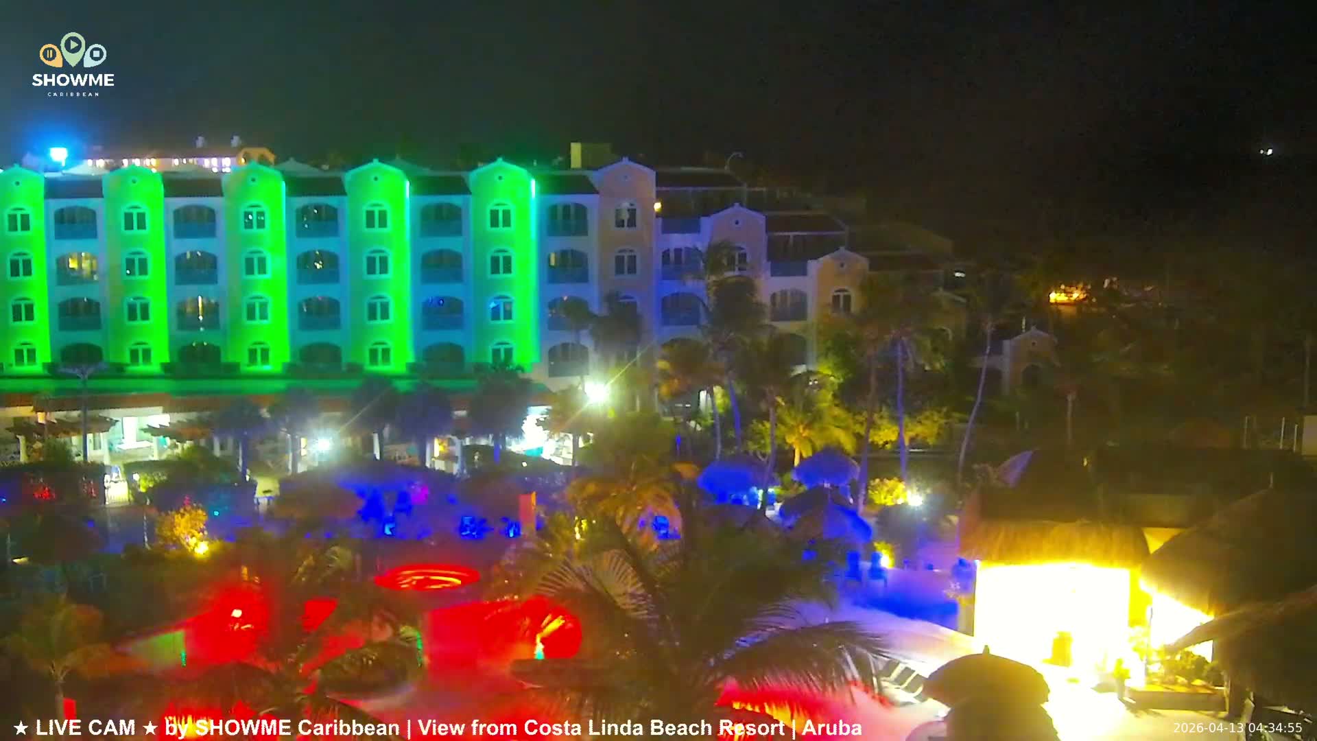 A vibrant resort complex is seen at night, featuring multi-story buildings illuminated in alternating green and blue lights, tropical palm trees, and sparkling blue swimming pools, all under a clear dark sky.