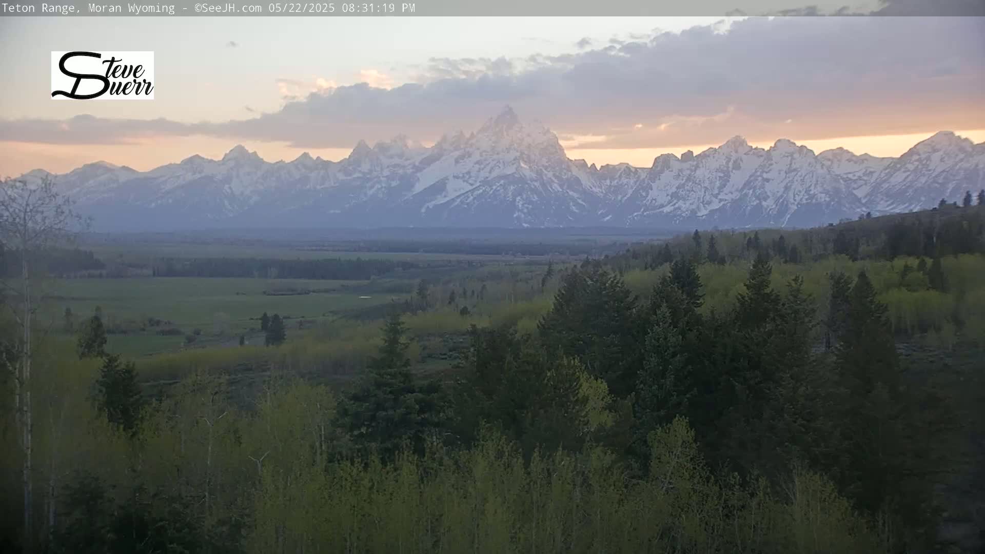 A snow-capped mountain range is visible at sunset, with a valley of green trees and fields in the foreground under a partly cloudy sky.