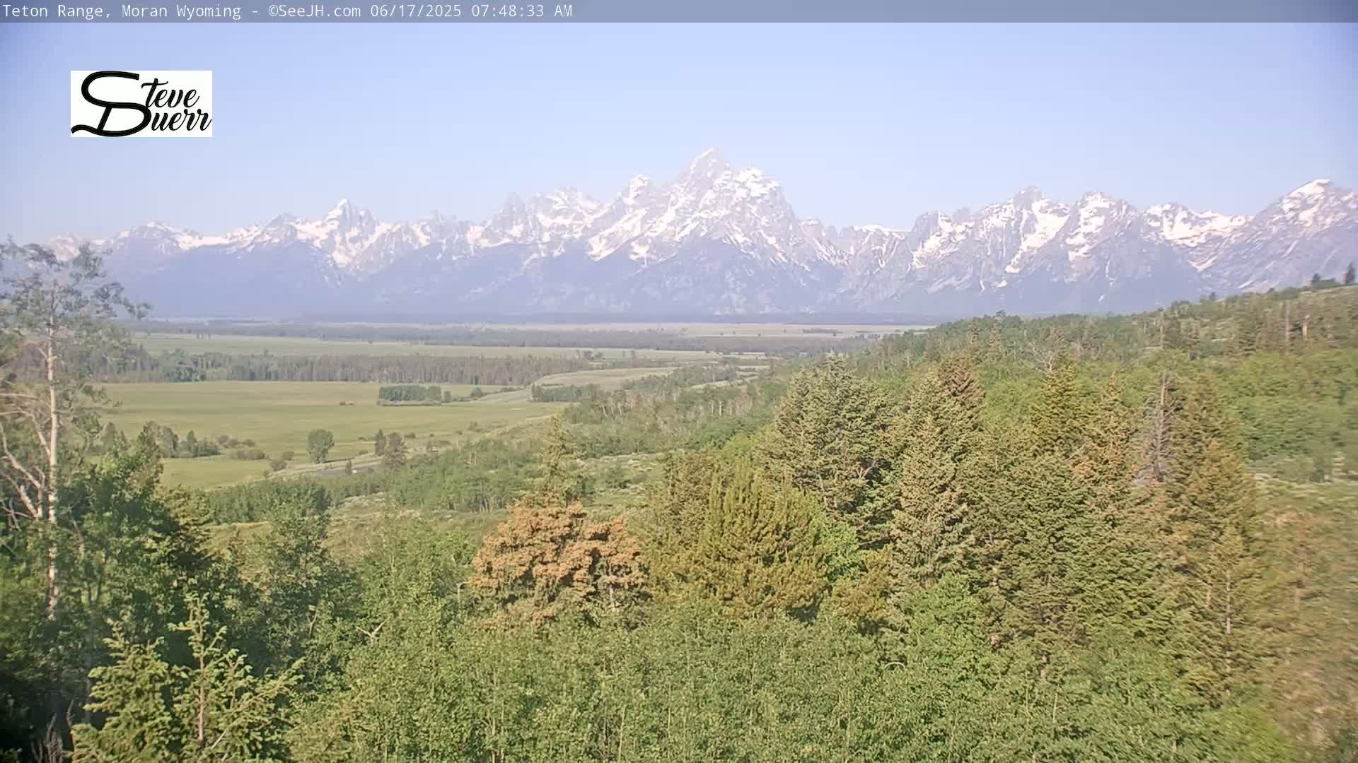 A panoramic view shows a snow-capped mountain range in the distance, beyond a valley of green fields and forests under a clear, bright sky.