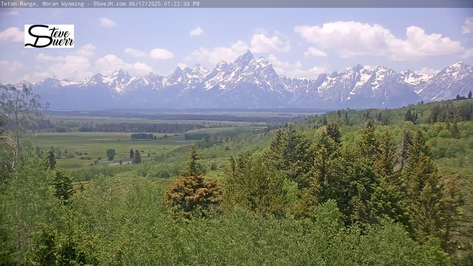 A partly cloudy sky overlooks a verdant valley and a snow-capped mountain range in the distance.