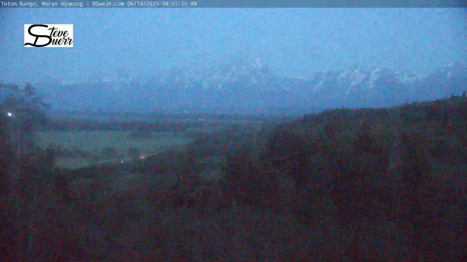 A hazy, dark-blue pre-dawn sky shows a distant mountain range, while the foreground is a shadowy landscape of trees and shrubs.