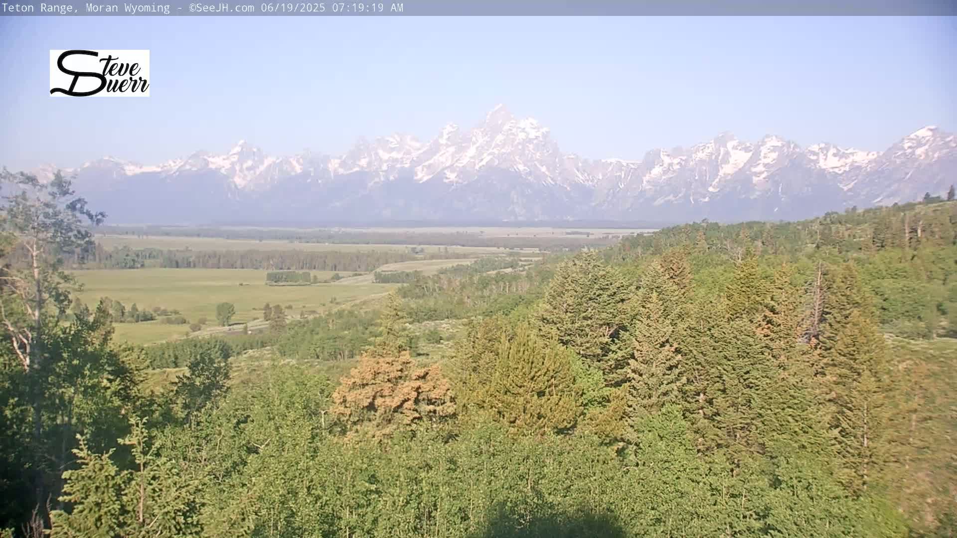 A snow-capped mountain range is visible in the distance beyond a valley of green trees and fields under a mostly clear sky.