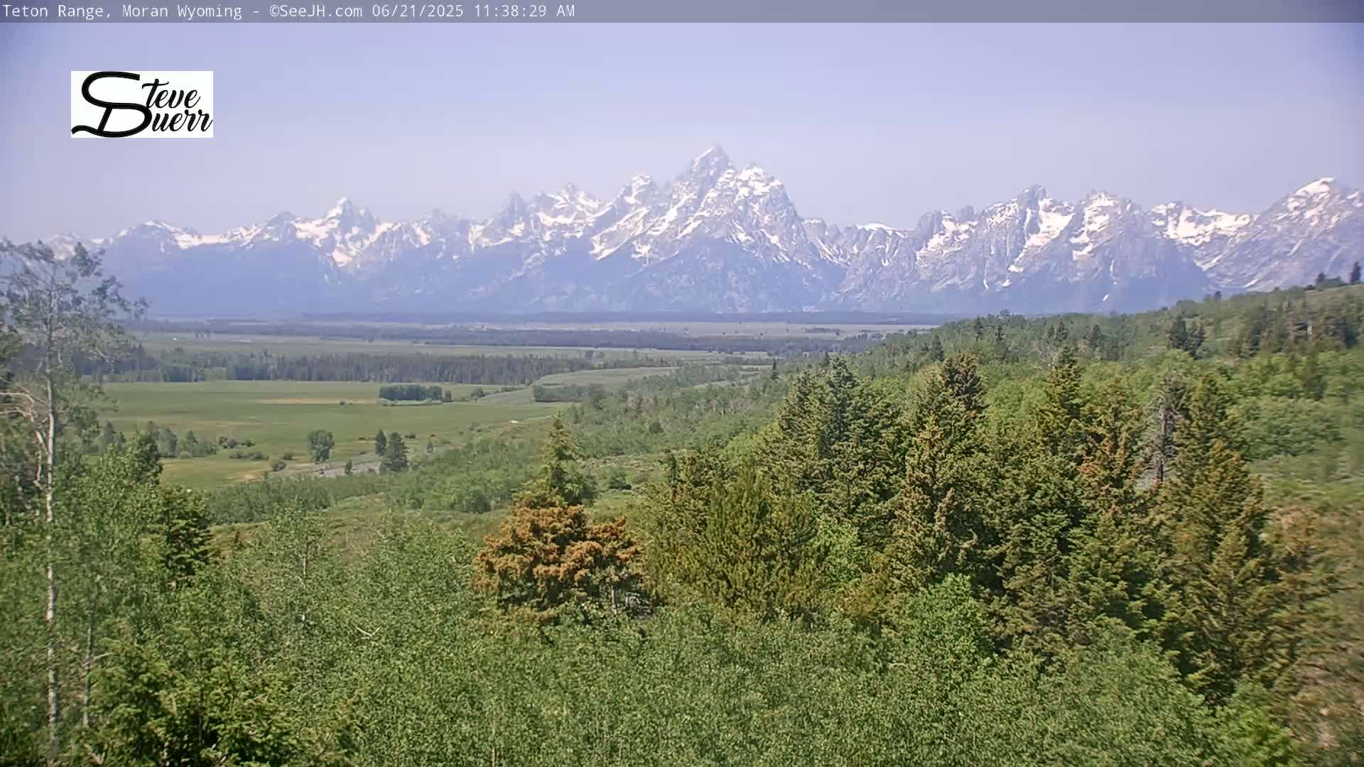 A snow-capped mountain range is visible in the distance behind a verdant valley and forest under a clear sky.