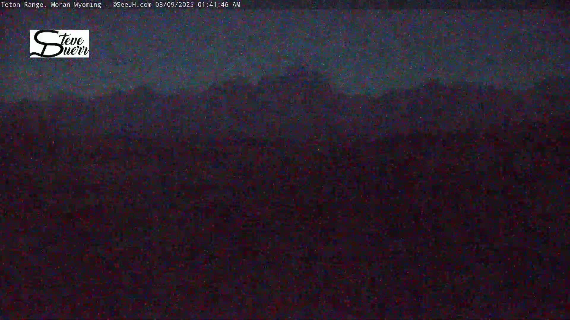 A dark, nighttime landscape shows silhouetted mountains under a mostly clear, dark sky.