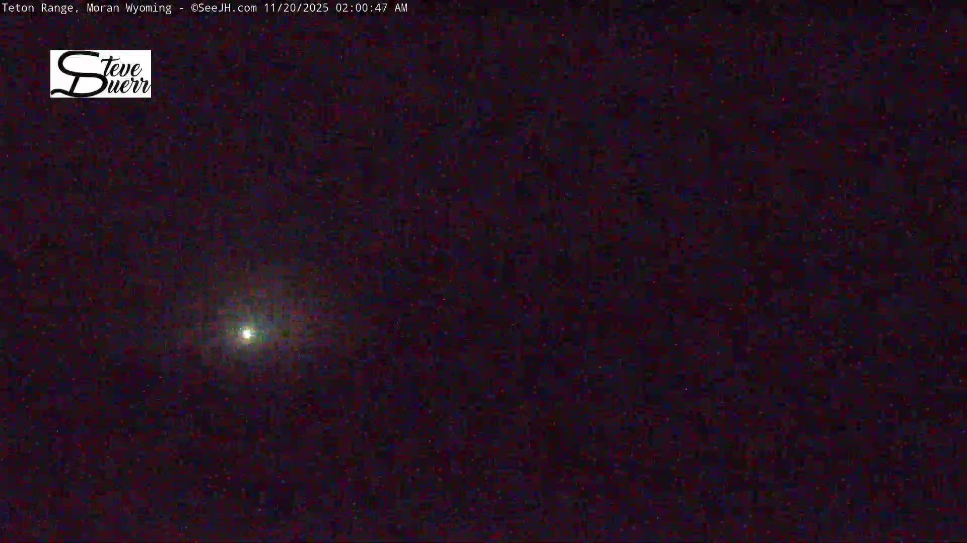 A dark, somewhat grainy night sky features a single prominent, blurry bright light source and numerous faint, scattered specks, under clear weather conditions.